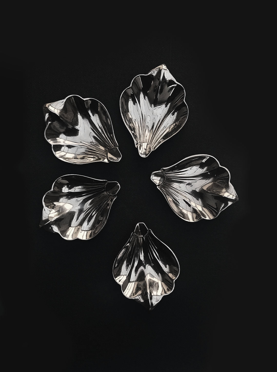 A set of five Petal Dishes from Les Objoies, each featuring a silver-plated leaf shape, are elegantly arranged in a circular pattern on a black background. The arrangement accentuates their shiny, reflective surfaces and intricate textures.