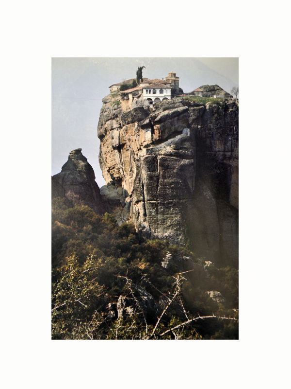 A monastery is perched on top of a towering rock formation amid a rugged, green landscape. The ancient building, rich in Greek history, is surrounded by cliffs and vegetation, standing alone and seemingly inaccessible, evoking a sense of tranquility and seclusion that can be further explored with Maison Plage's "The Greek World: Classical, Byzantine, and Modern.