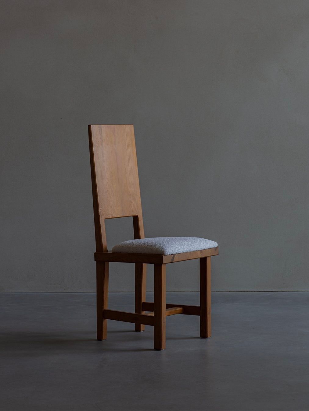 A Göran Malmvall - Model 553 chair by Bicci de' Medici, featuring a wooden frame, tall rectangular backrest with a square cut-out, and light gray padded seat, stands against a dark gray backdrop, capturing the essence of Swedish minimalist design.