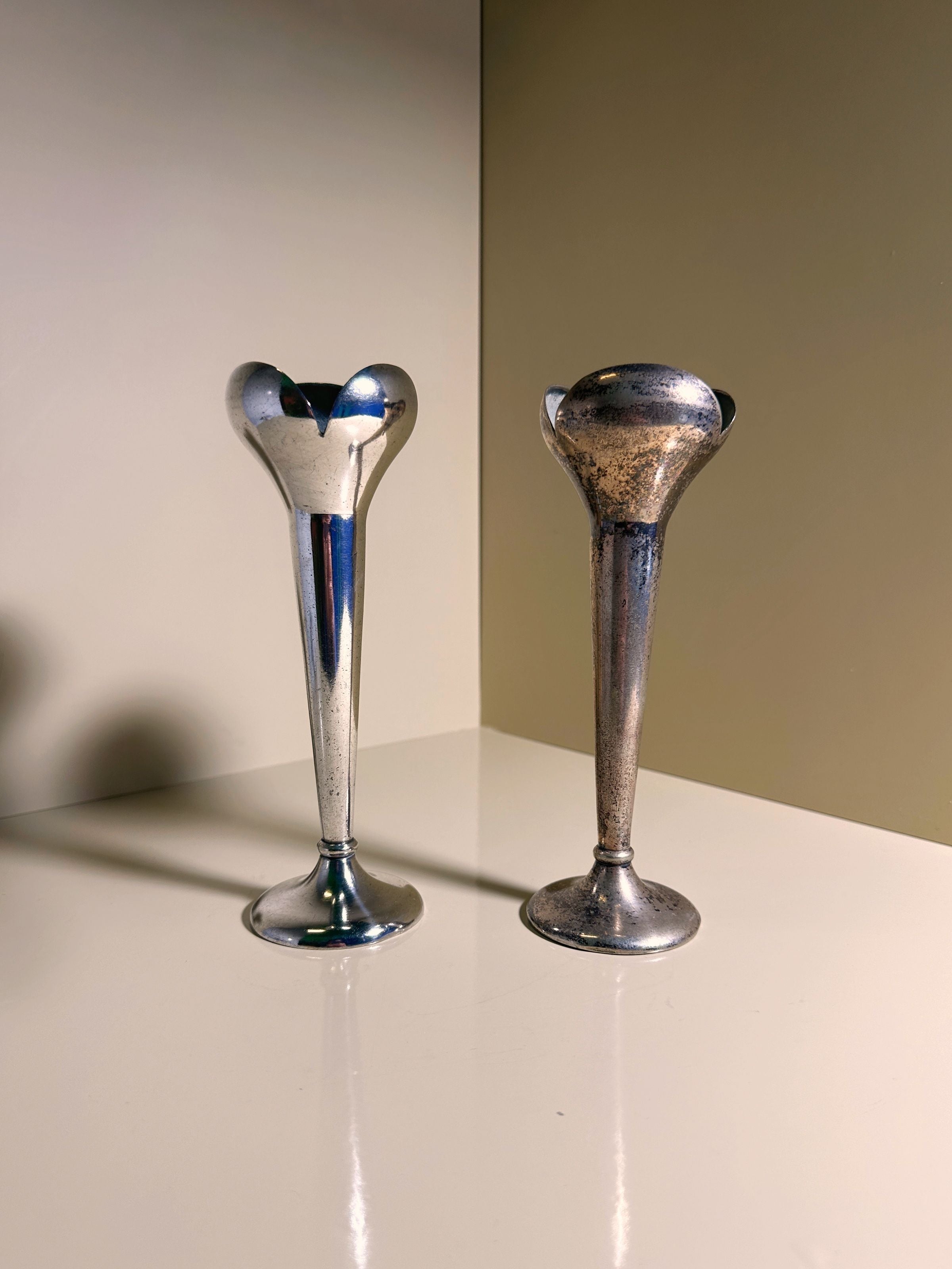 A set of two Gio Ponti candlesticks by Calderoni from the 1930s, one gleaming in Alpacca silver and the other charmingly tarnished, rests on a light surface. These Dodo Vintage pieces sit against a neutral backdrop with subtle shadows.