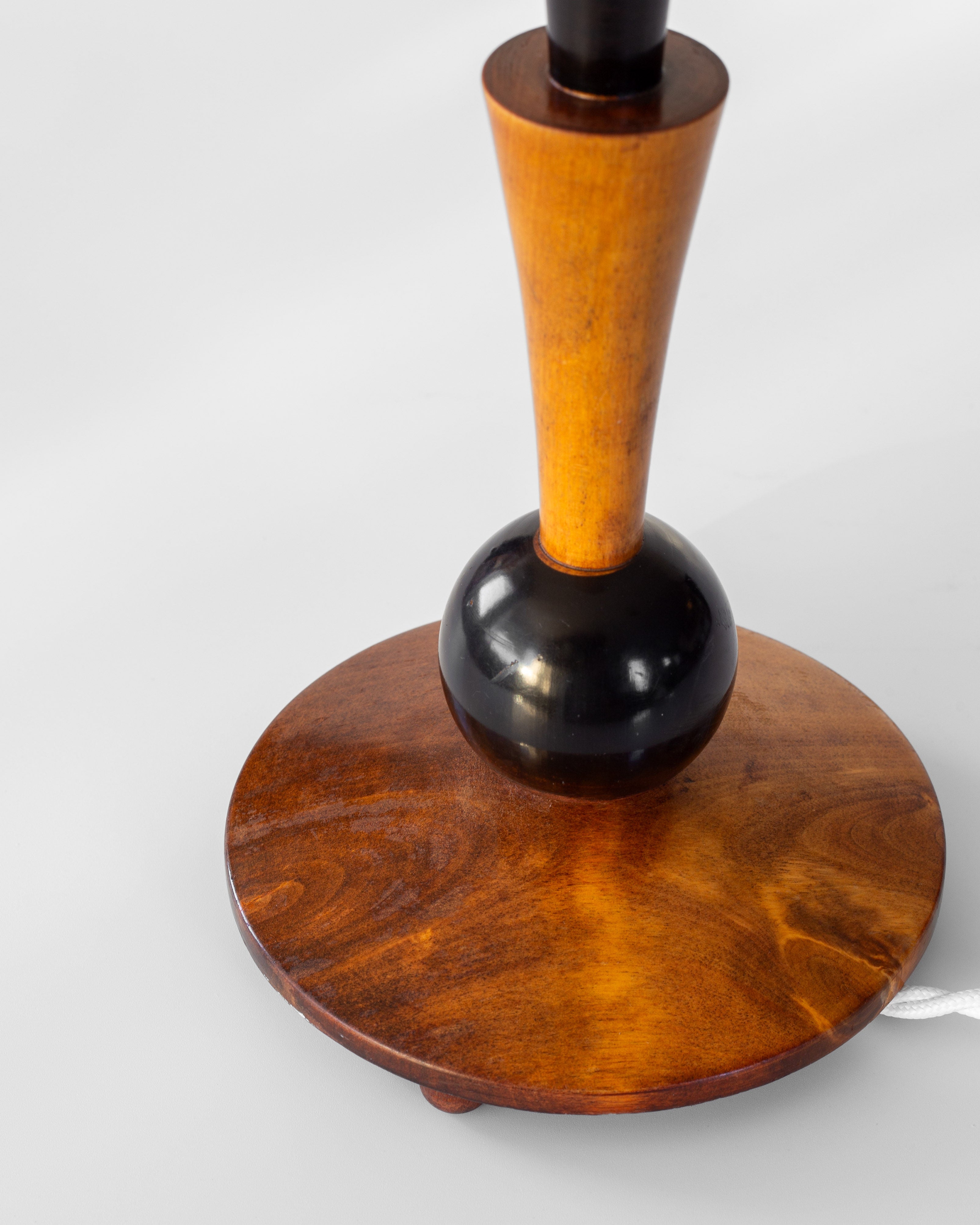 The Spigel Art Deco Wooden Table Lamp showcases a vintage charm with its circular, polished surface and a tall, narrow neck. It includes a dark, spherical accent near the center reminiscent of an Art Deco design, set against a light gray backdrop. A white cord is elegantly integrated from beneath the base.