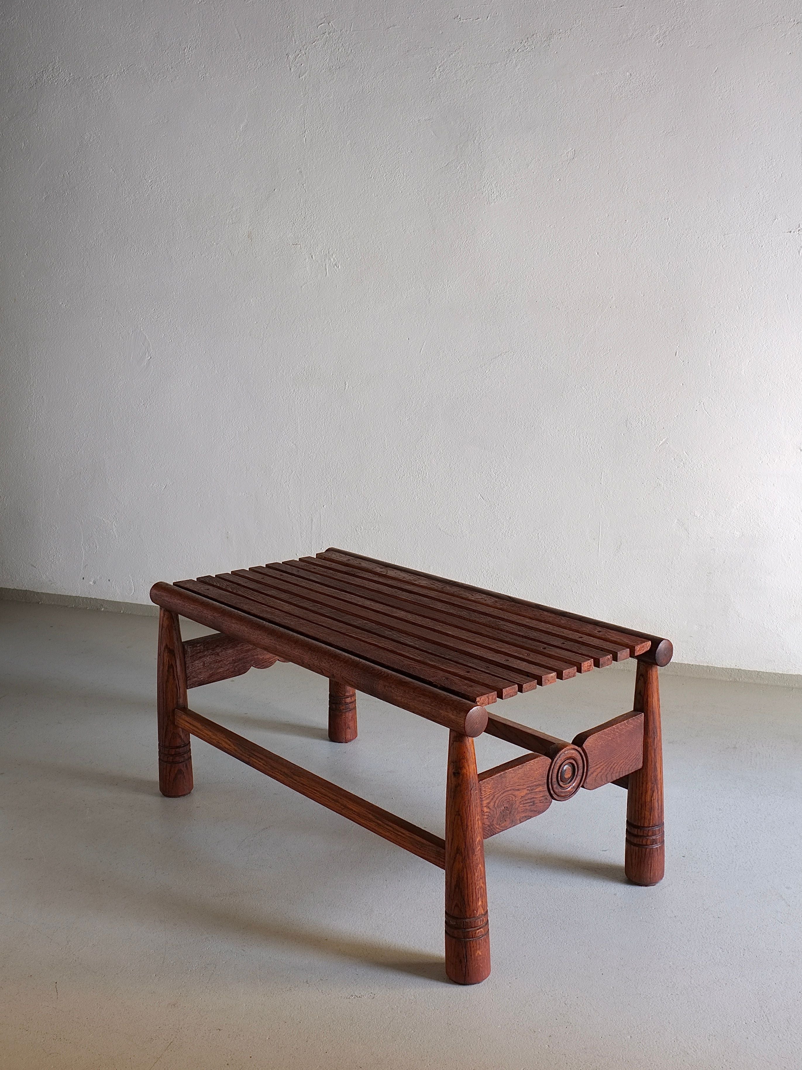 The Carved Dark Oak Bench by Veter Vintage, a vintage piece from France dating back to the 1960s, stands against a plain, light gray wall. This small bench features a slatted seat and decorative circular designs on the legs. The dark-stained oak showcases visible wood grain and minimal ornamentation for a simple, rustic aesthetic.