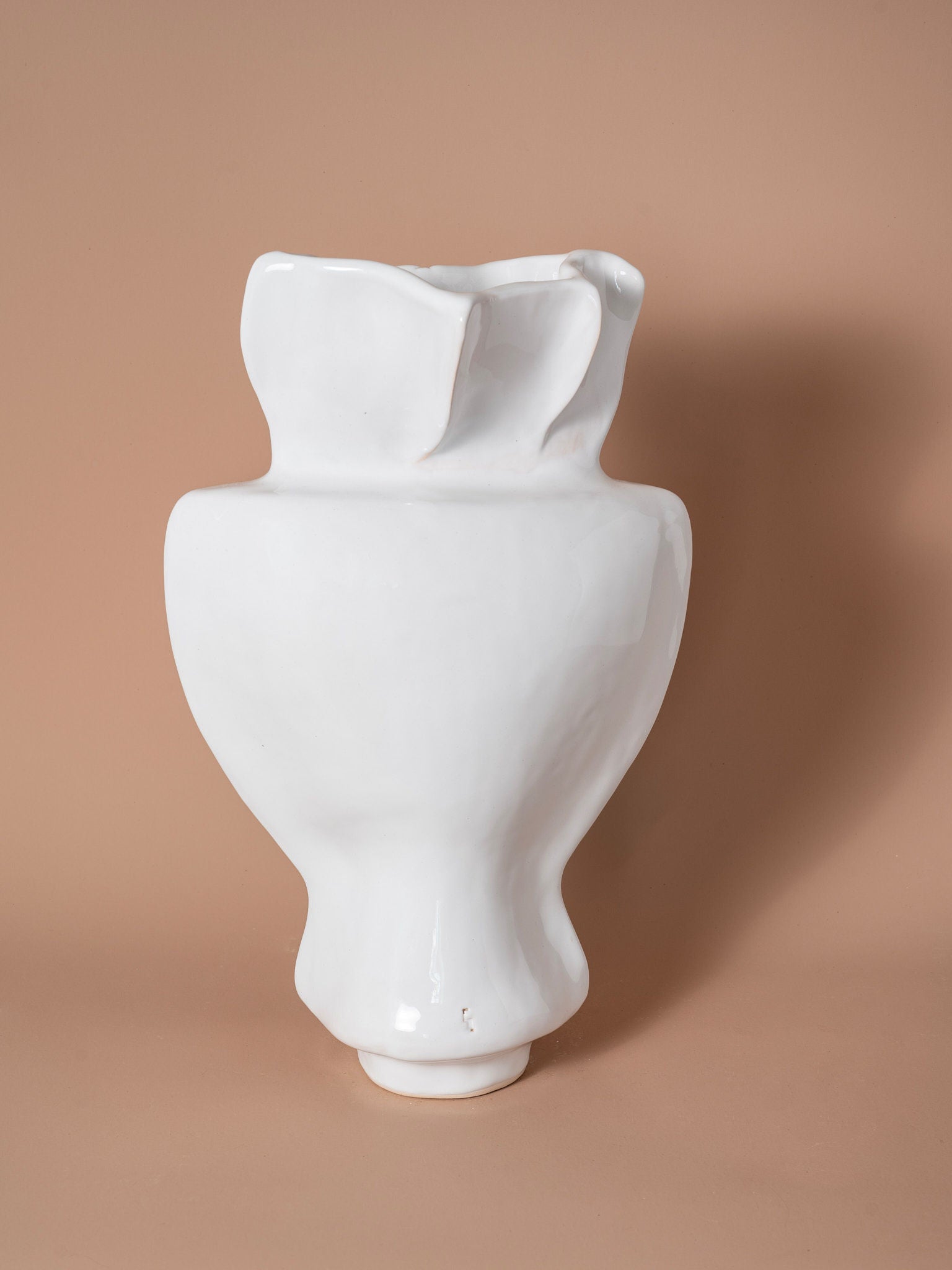 The Seed of Light Form Vase 14 from Siham Djebbar Ceramics’ Seed Series is a white, glossy handcrafted stoneware vase with an abstract shape, wide base, narrow middle, and irregular rim set against a light brown background.