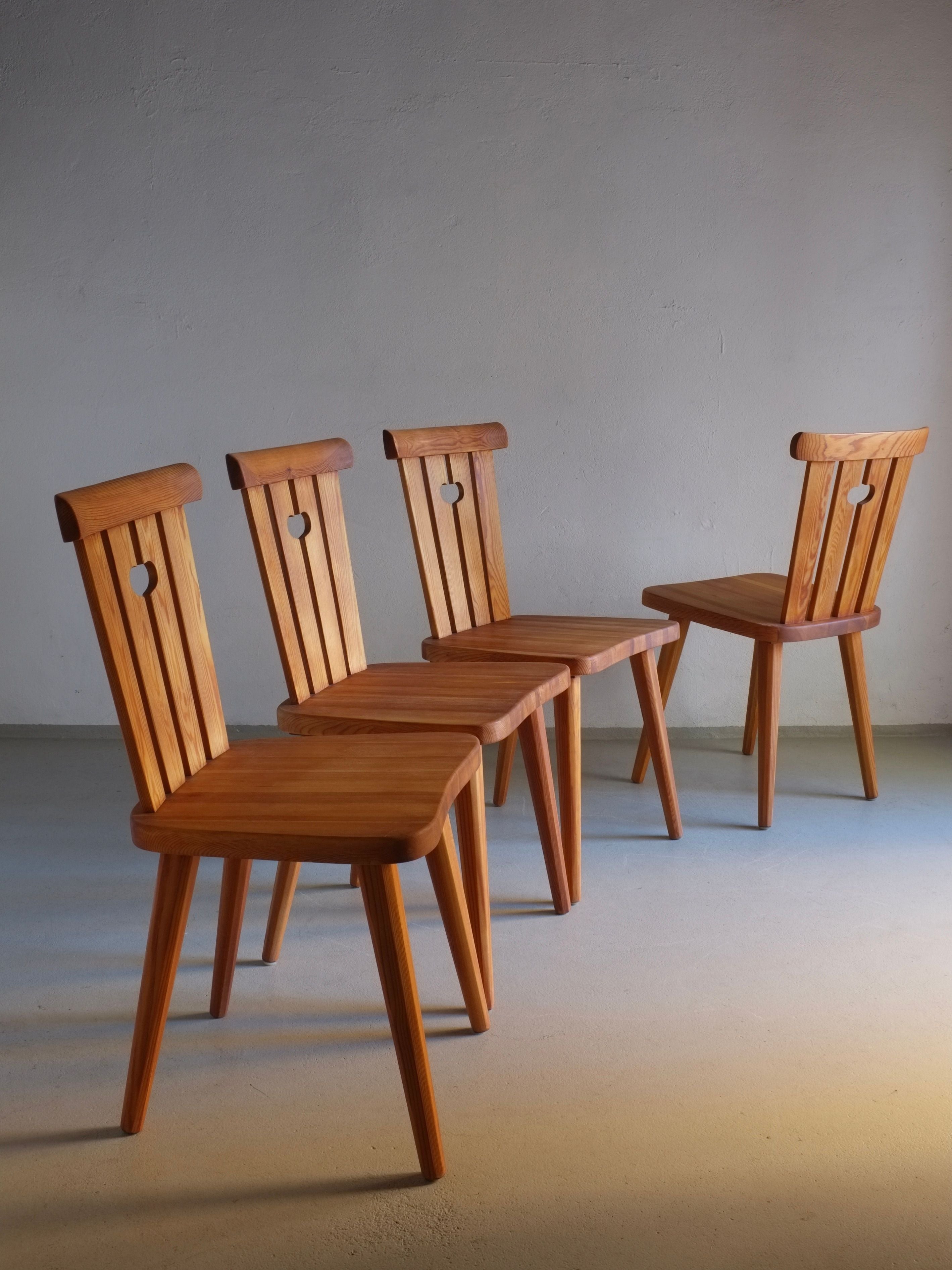 Four rustic pine chairs by Göran Malmvall from the 1940s, offered by Veter Vintage, are arranged in a staggered line. Each chair features a heart cutout in the backrest, standing proudly on a light-colored floor against a plain gray wall. The lighting casts soft shadows on the floor.