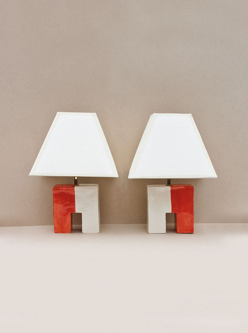 Two hand-built Cristo Lamps (Rojo/Hueso) by Casa Veronica with custom square shades feature unique bases: the left side of each base is white and the right side is red. The bases showcase a symmetrical, geometric design with a rectangular cutout in the middle. UL listed for safety, these elegant lamps stand against a plain, neutral-toned background.