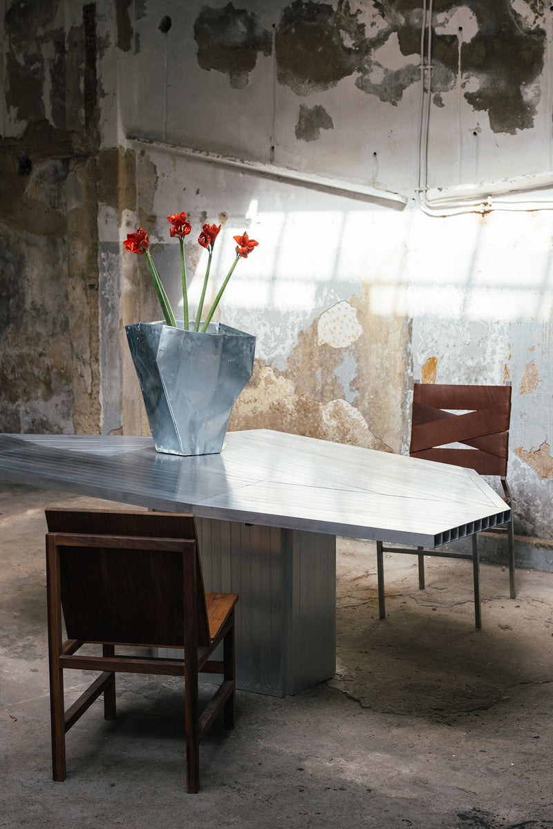 An Aluminum Dining Table by Studio HAOS stands in a weathered industrial room. A geometric vase with red flowers sits on top, flanked by two mismatched chairs, reflecting the brand’s signature contemporary design.