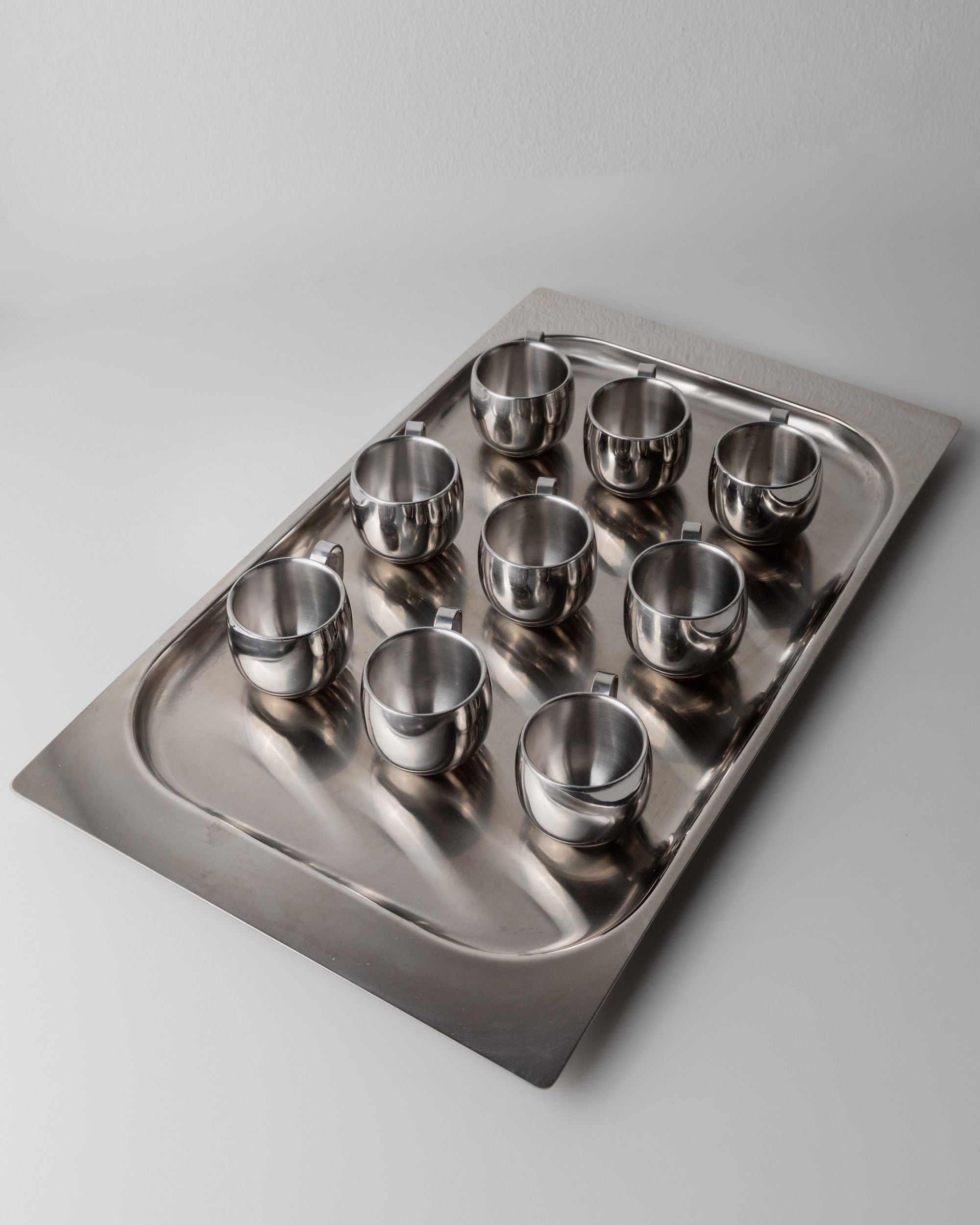 A silver tray showcases the vintage "Coffee set by IMCO 70s" from Bottega Jacobs, featuring ten elegantly aligned stainless steel cups with handles, all perfectly arranged on a plain, light-colored surface. This display embodies minimalist design at its finest.