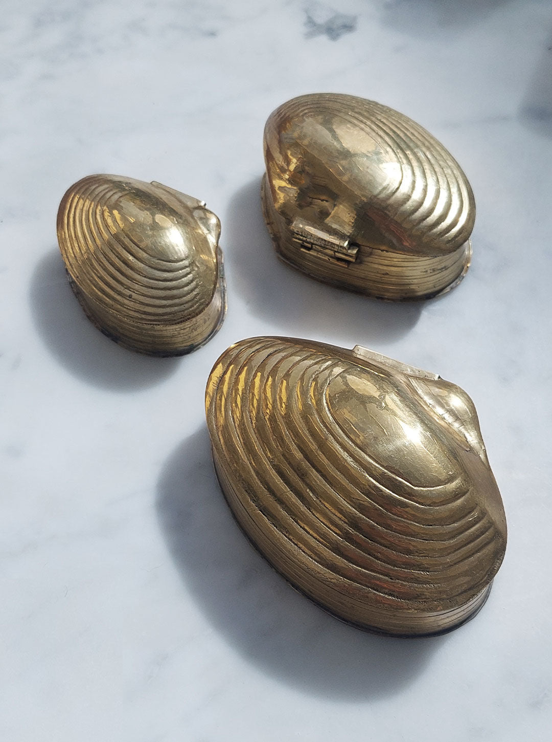 Three vintage Les Objoies Set of 3 Nesting Shell Boxes with a brass gold finish are displayed on a white marble surface. The largest box is positioned at the top, while the smaller two are arranged below it. The boxes feature hinged lids and detailed shell ridges, creating elegant nesting boxes.