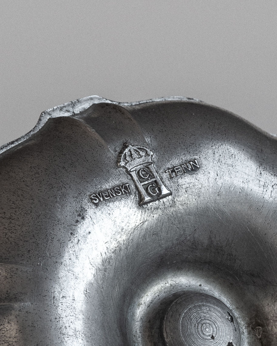 Close-up of a Modernist Metal Candleholder by Spigel from the 1930s. The candleholder features an embossed emblem with a crown-like symbol above the letters "CG" and the text "SVENSKT TENN" inscribed below, reminiscent of Swedish Modernist metalwork by C.G. Hallberg. The surface boasts a worn, textured finish.