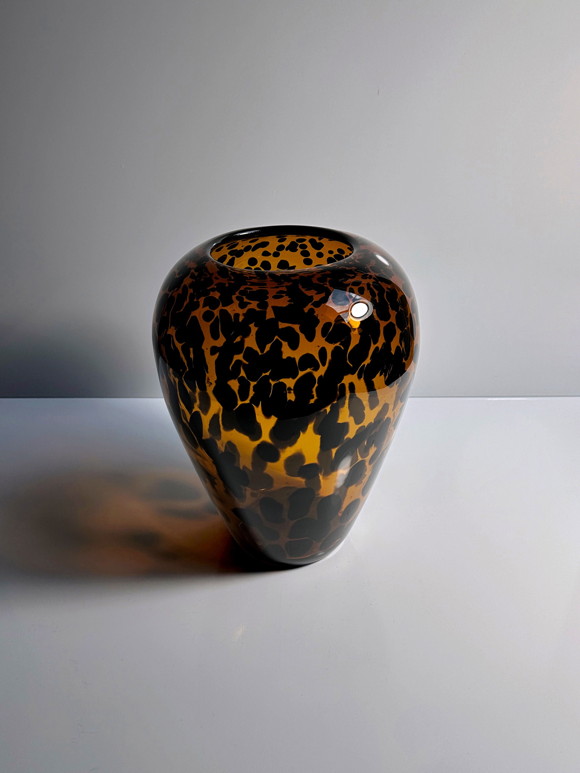 A glossy, spherical Tortoise vase Murano for Christian Dior from the 1960s, available through Dodo Vintage, features a leopard print pattern in shades of brown and black. It is elegantly displayed on a white surface against a gray background, beautifully reflecting light on its finish.