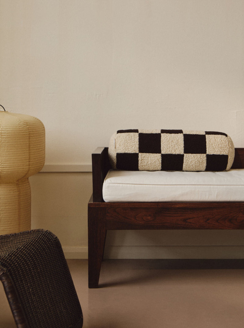 A minimalist interior features a wooden bench with a vintage-inspired cushion and a black and white checkered Le Cylindre - Wool Bouclé - Chocolate/Eggshell bolster pillow from AMÉLIORÉ. To the left, there is a textured, round beige lamp handmade in the UK. In the foreground, part of a woven chair is visible. The background has a plain, light-colored wall.