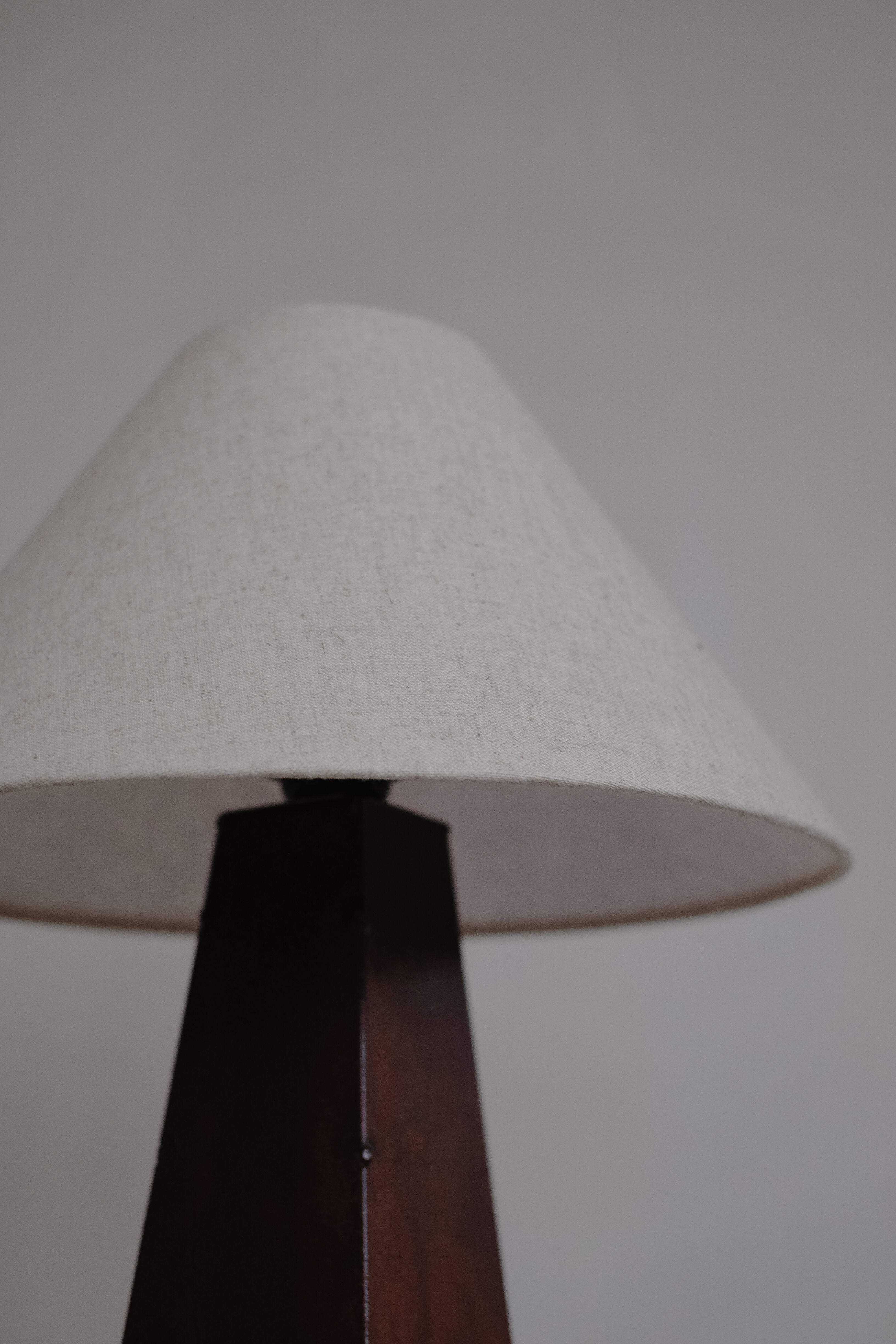 A close-up of the septembre studios Obelisk Table Lamp, showcasing a beige fabric lampshade and dark wooden base on a gray background. The image emphasizes the lamp's elegant design, texture, simplicity, and dimmable switch for customizable lighting.