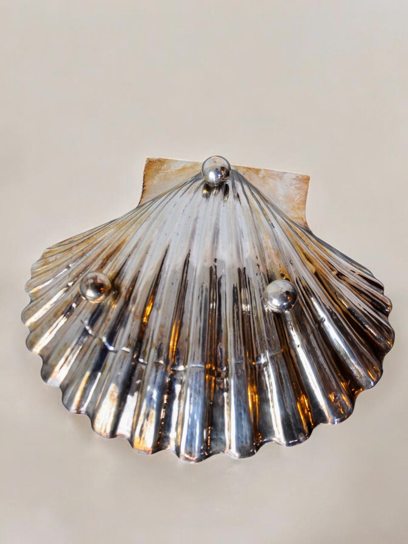 The Shell Vide-poches by Malamar Studio, a sleek metallic scallop shell from the 70s Valenti collection, showcases three small metal spheres on its surface, set against a plain background.