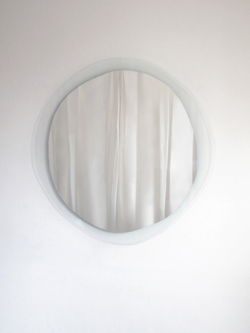 A round Mirror Ombrée Clair from Laurène Guarneri with a wavy edge hangs against a plain white wall. The reflection shows a light-colored, possibly white, curtain with gentle folds. The minimalist setting emphasizes the simplicity and elegance of the handmade mirror's unique composition.