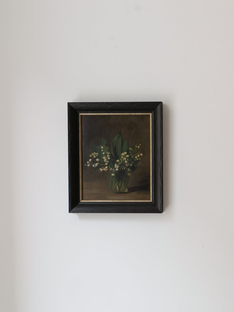 A framed Lily of the Valley Still Life 1961 painting by Collection apart hangs on a plain white wall. The dark frame contrasts with the light background, highlighting the delicate white blooms and green leaves in their green vase, reminiscent of Dutch still life art.