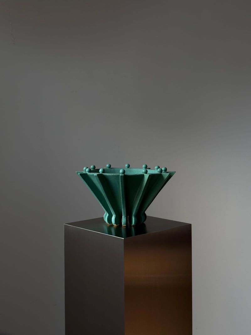 Vessel No. II by Devin Wilde is a sculptural green ceramic bowl with angled sides and small round knobs on the rim, displayed on a reflective square pedestal against a plain gray background.