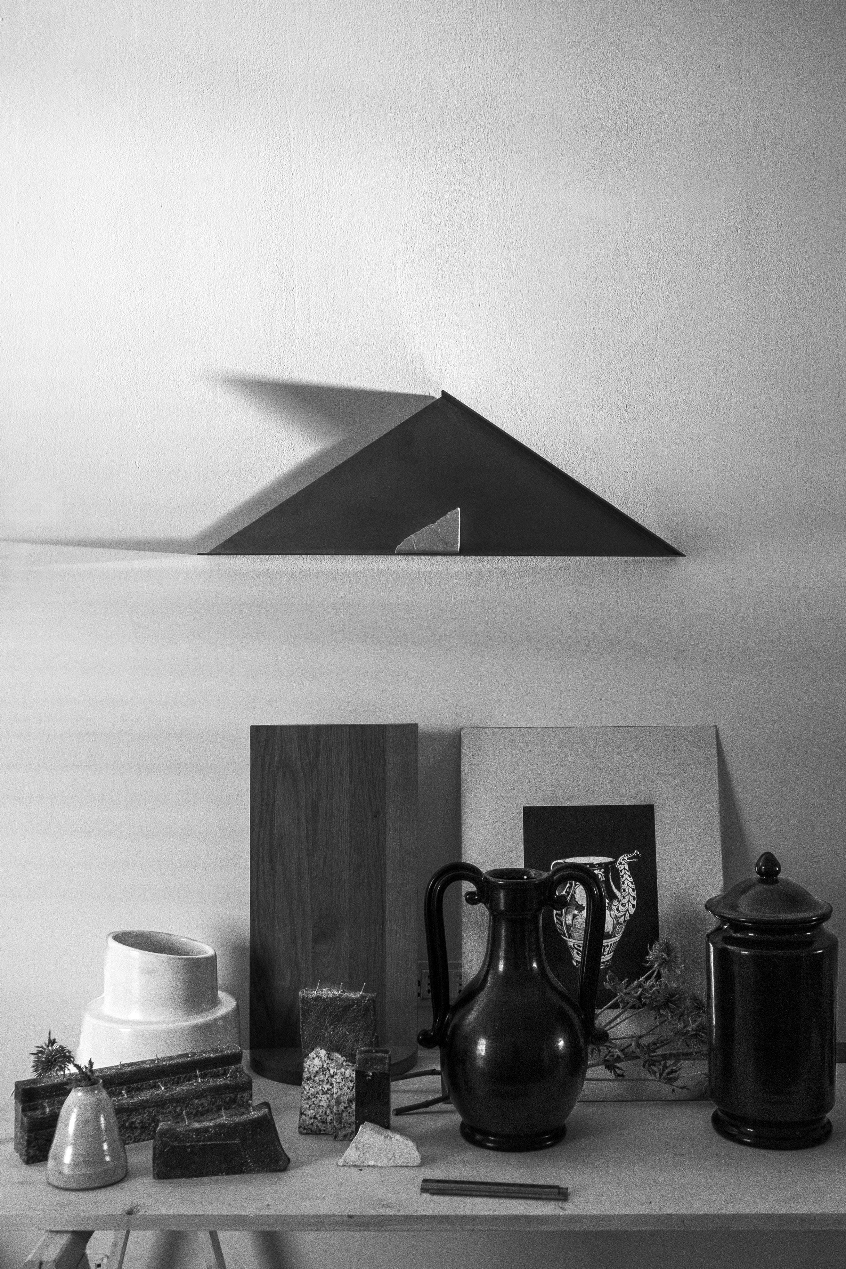 A black and white image of a tabletop with jars, bowls, plants, textured blocks, and framed artwork arranged before a white wall with the limited edition COSEINCORSO "The Front - Shelf in Metal" mounted above.