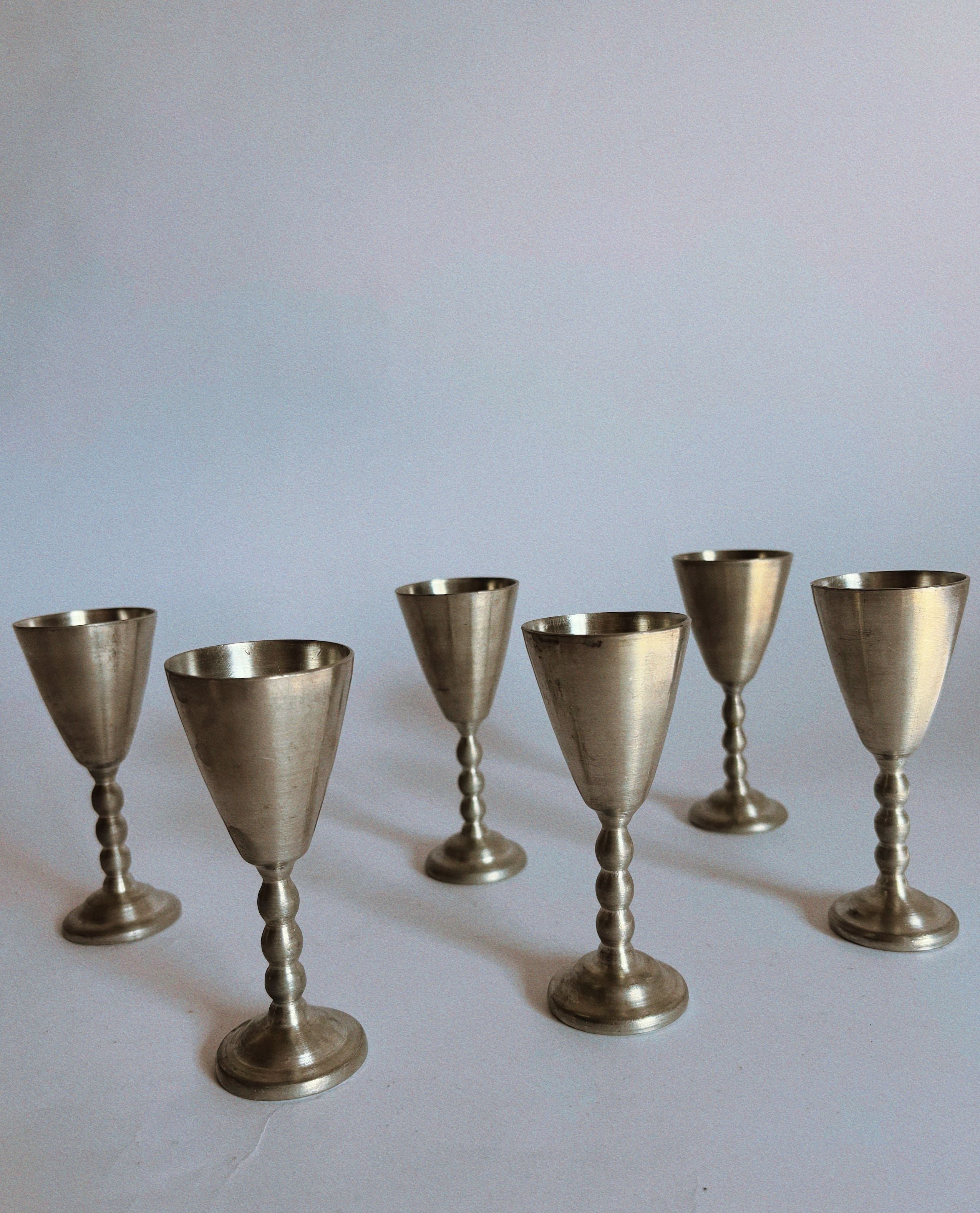 The Huerto Set of 6 Pewter Schnapps Goblets from Sweden features vintage goblets with a matte finish, slender tiered stems, and rounded bases, elegantly arranged on a light-colored surface against a neutral background.