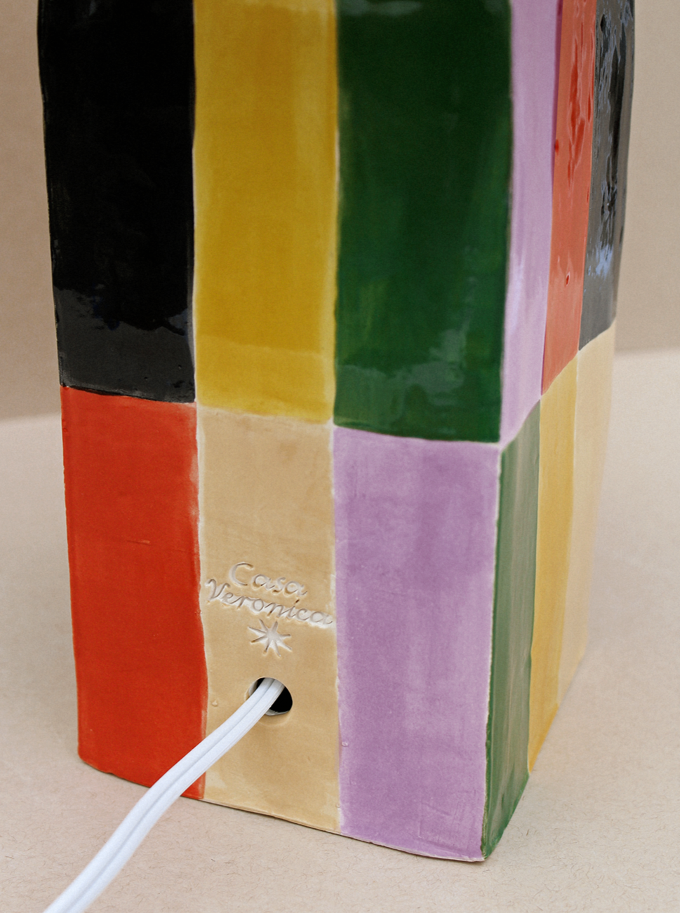 A colorful Casa Veronica Milagro Lamp (Grande) with vertical stripes in black, yellow, orange, green, and purple, featuring an E26 base and plugged into a wall socket by a white cable.