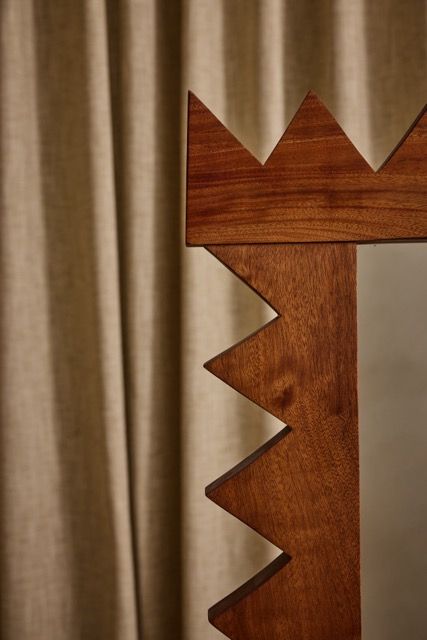 Close-up of the Jura Mirror by Ah Um Design Studio, showcasing its bold solid mahogany frame with zigzag crown-like edges, set against beige curtains in the background.