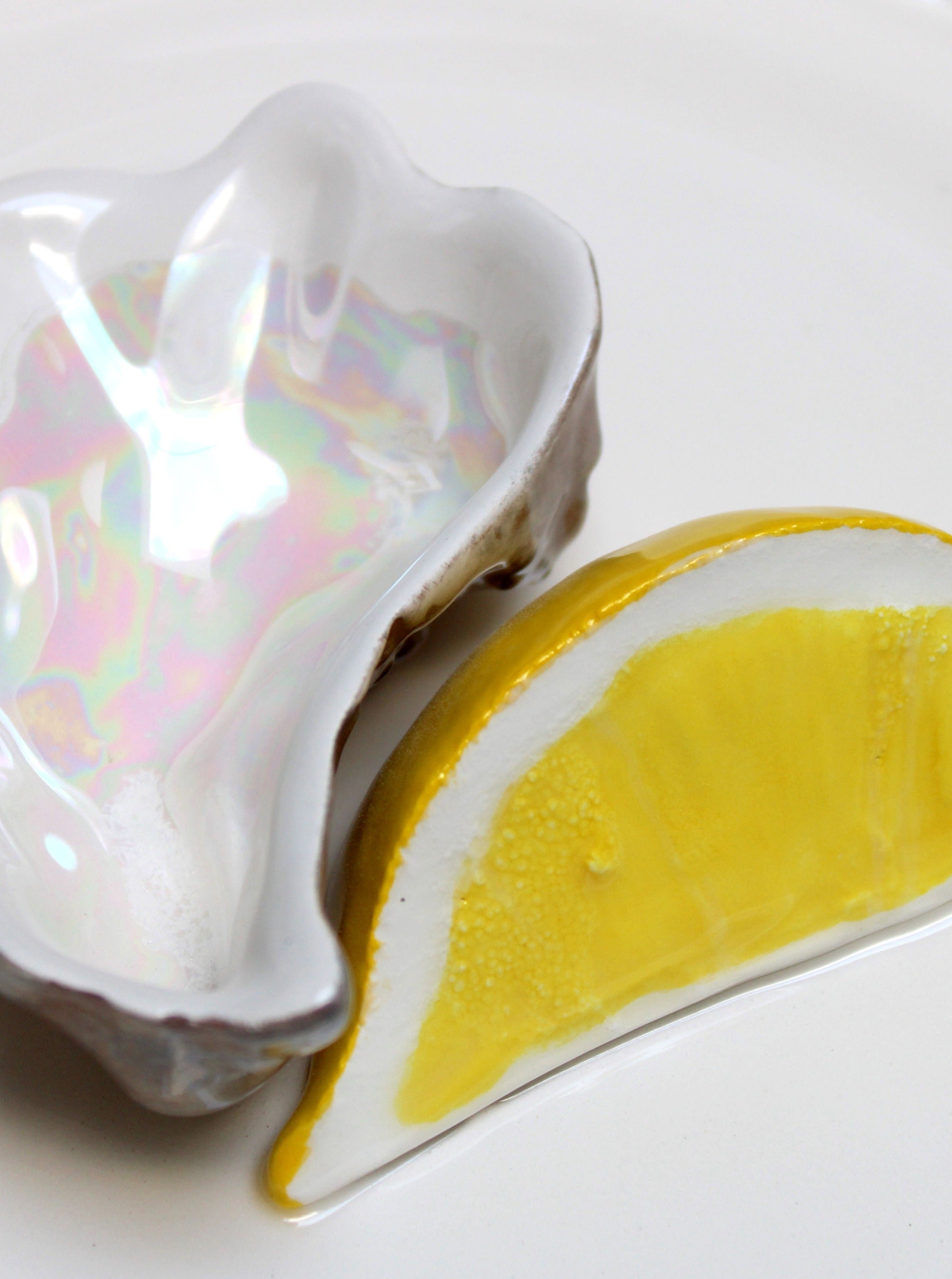 The Plaisir Solitaire Ashtray by Villa Arev, featuring an iridescent ceramic oyster shell design, is paired with a ceramic lemon wedge on a white plate for a sophisticated touch.