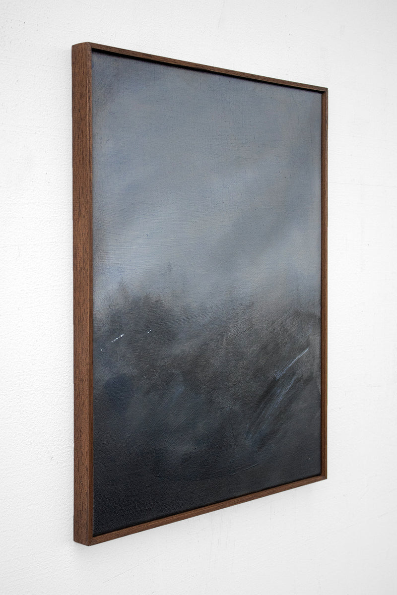 As Night Draws In by David Hardy is an original abstract acrylic on linen, featuring dark, smoky tones of black, gray, and blue that evoke a moody landscape. Framed and displayed on a white wall, it’s viewed from an angle.