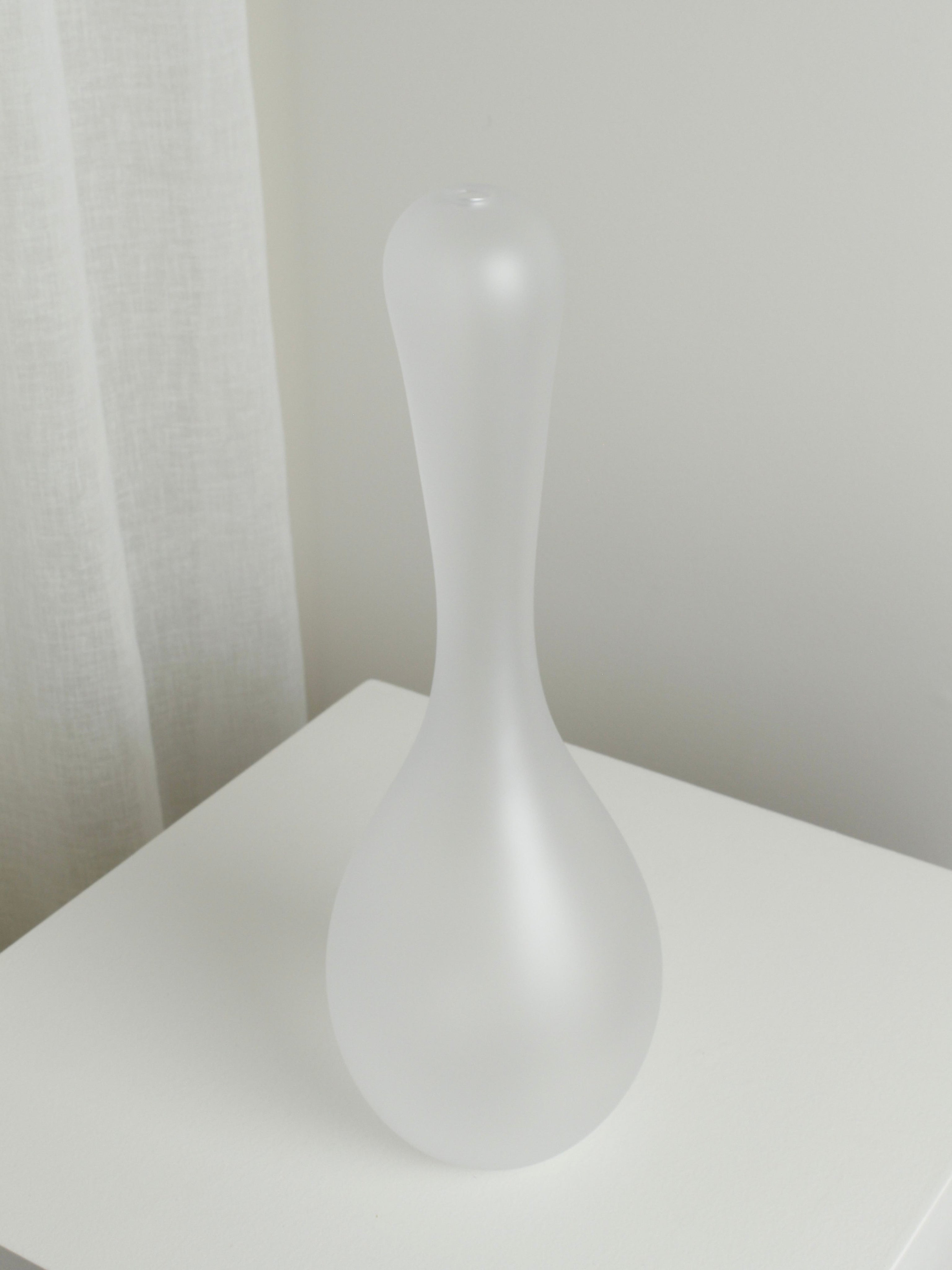 The XLarge Vase in Ghost by Verre d'Onge, featuring a narrow neck and bulbous base, rests on a white surface near a sheer curtain and light wall, bringing an ethereal touch to any space.