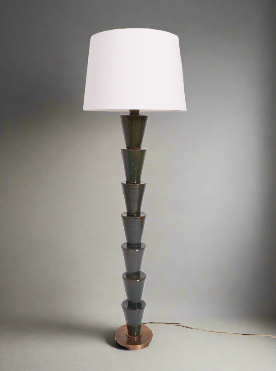 The "Nizwa" Floor Lamp by Barracuda Interiors features a stacked, geometric terracotta base with a round wooden bottom and a large white cylindrical shade. It offers customizable lighting and has a visible floor cord.