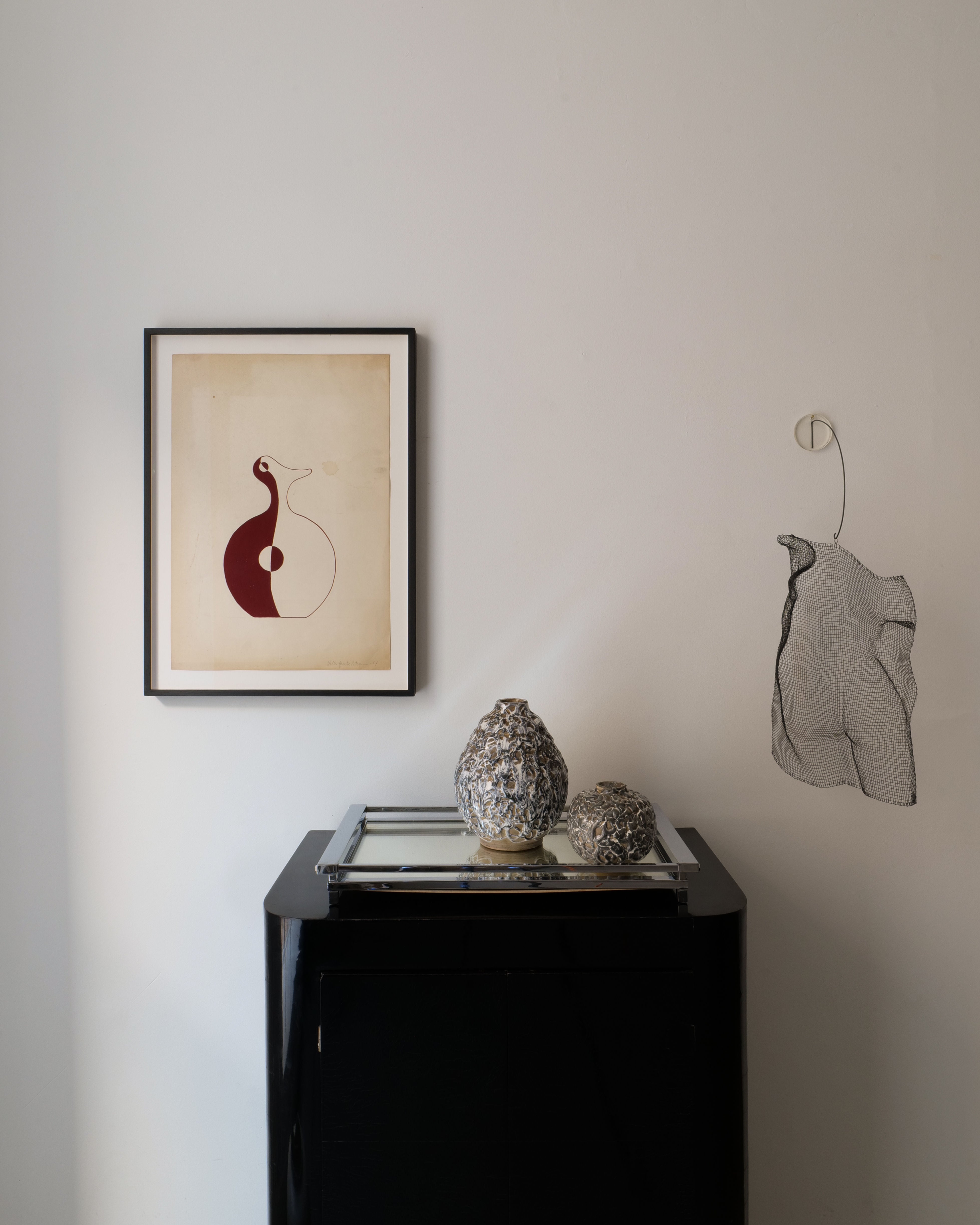A minimalist interior featuring a black cabinet, two textured sculptures, a Collection apart 'Red Vase' Painting 1951 framed on the wall, and a wire mesh sculpture—all set against a plain white background.