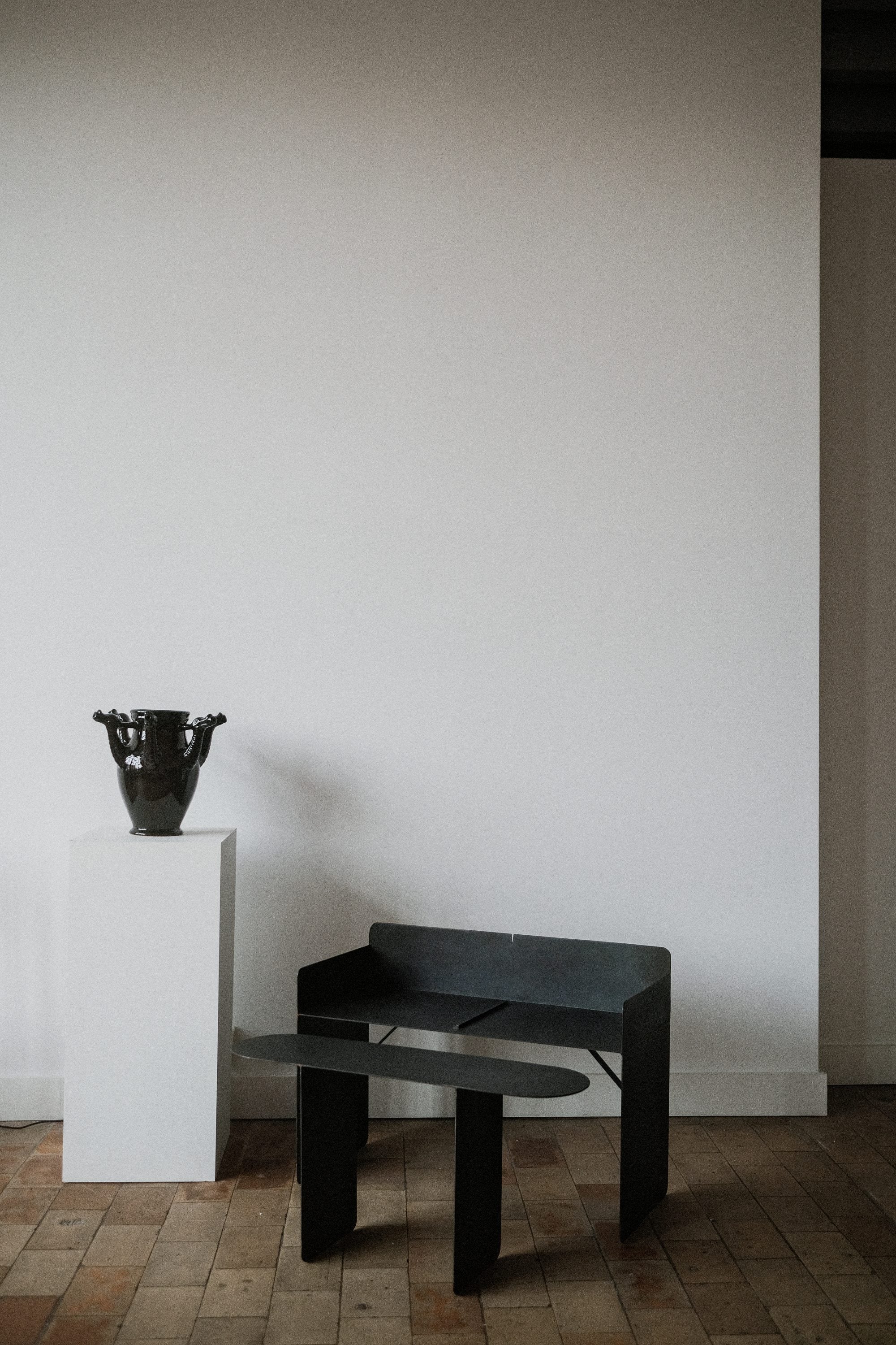 A minimalist interior showcases a dark metallic bench and The Drago Bianco - Minimalist Vase in Ceramic by COSEINCORSO on a white pedestal, set against a blank white wall and wooden floor.