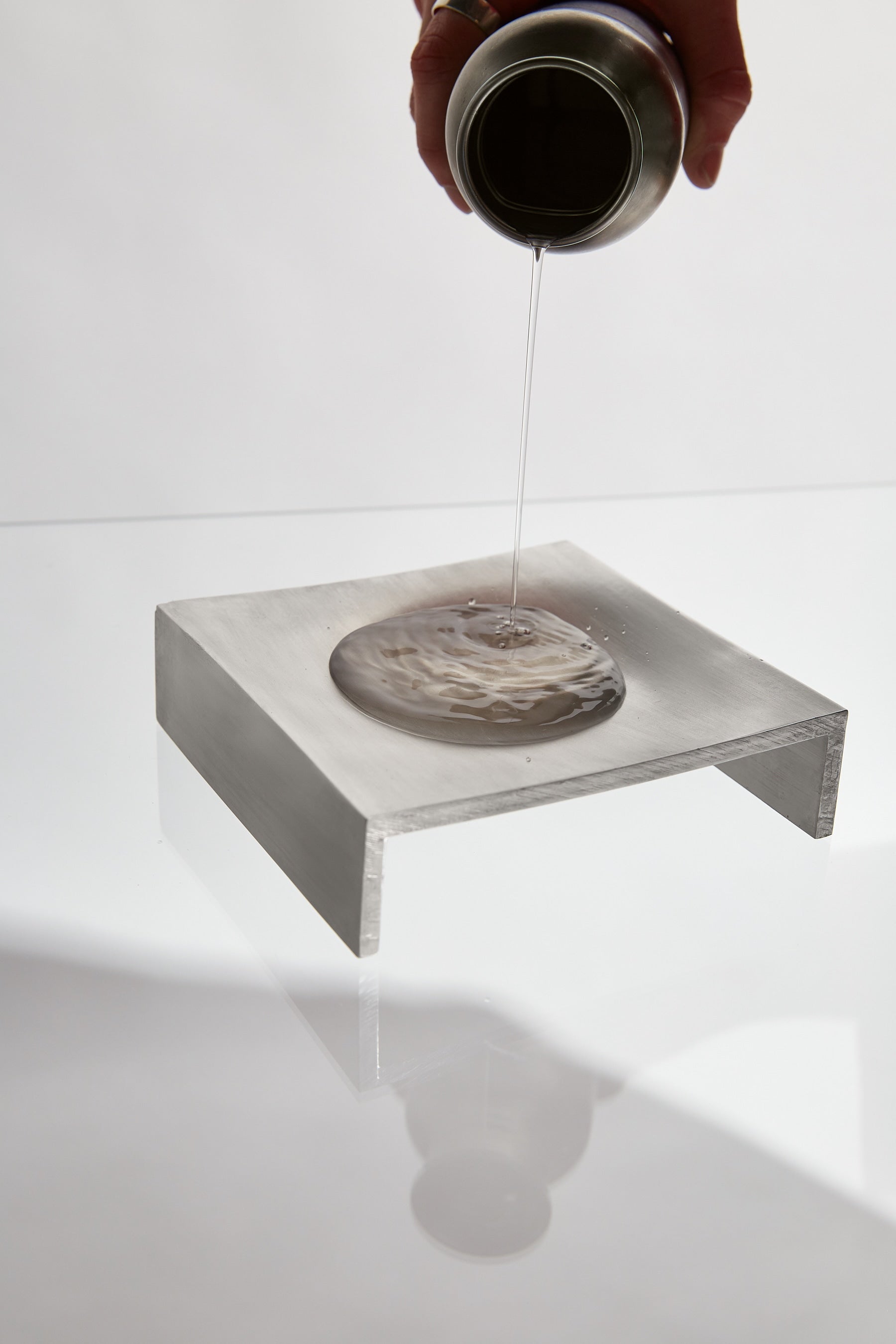 A hand pours a clear liquid from a metal container onto the LESORR Square Aluminum Vide-Poche, which rests on a rectangular aluminum platform against a white background.