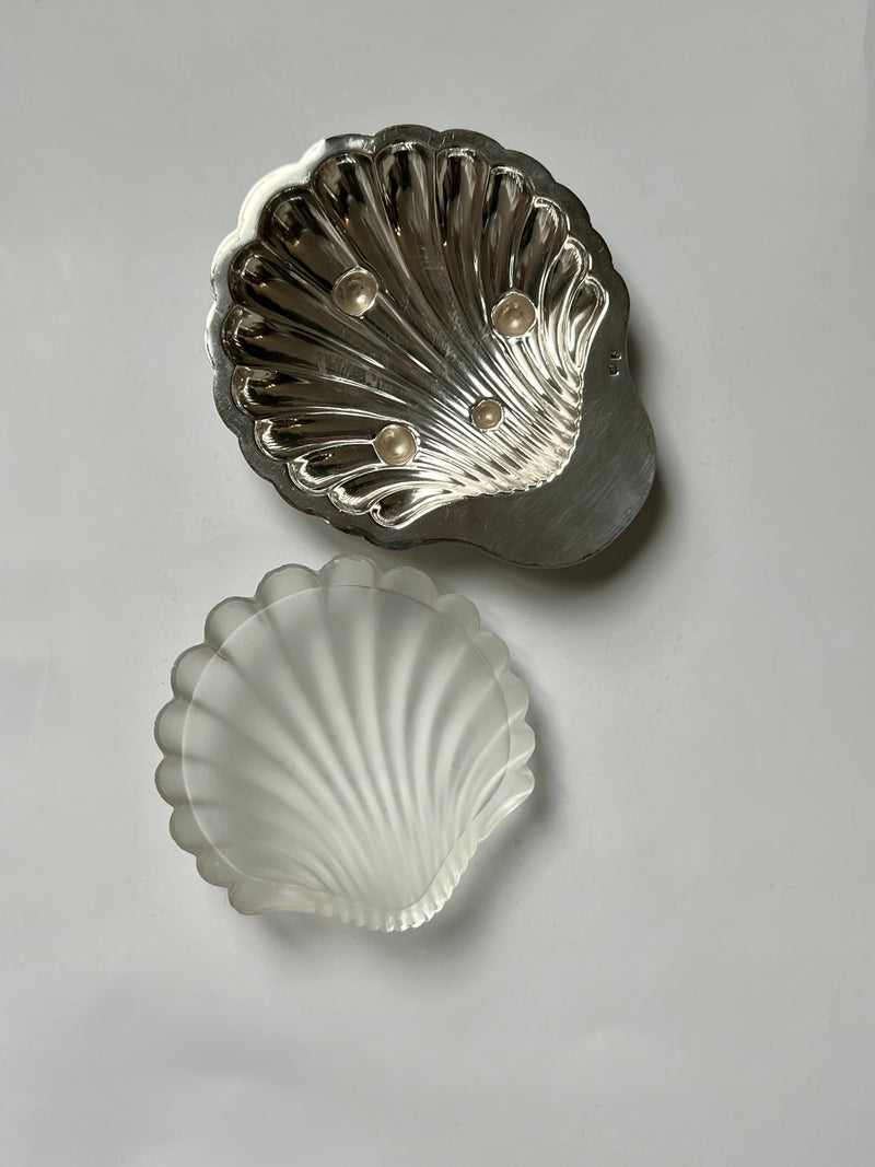 Two shell-shaped dishes are displayed on a white background: the top is the Manushi Silver Shell Shaped Dish holding three small pearls, while the bottom dish is translucent and frosted.