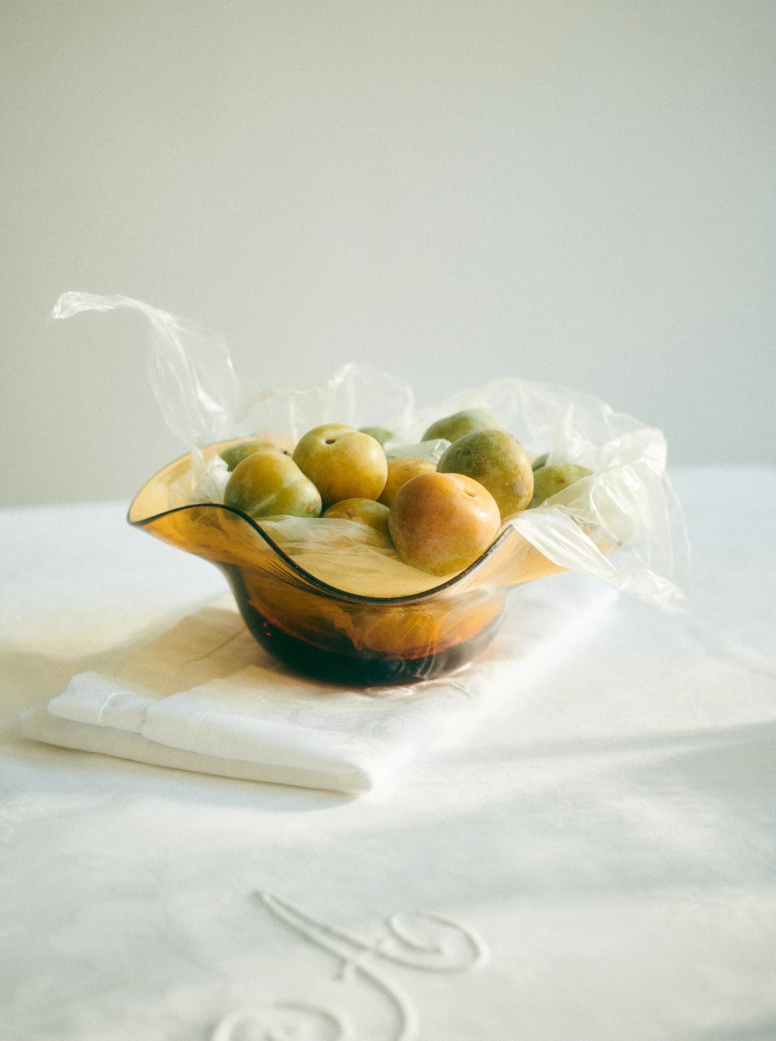 The Michele Bowl Small by Akua Objects, filled with green and yellow plums and partially covered in translucent plastic, rests on a white cloth-draped table. Soft natural light highlights the bowl in a serene, minimalist setting.