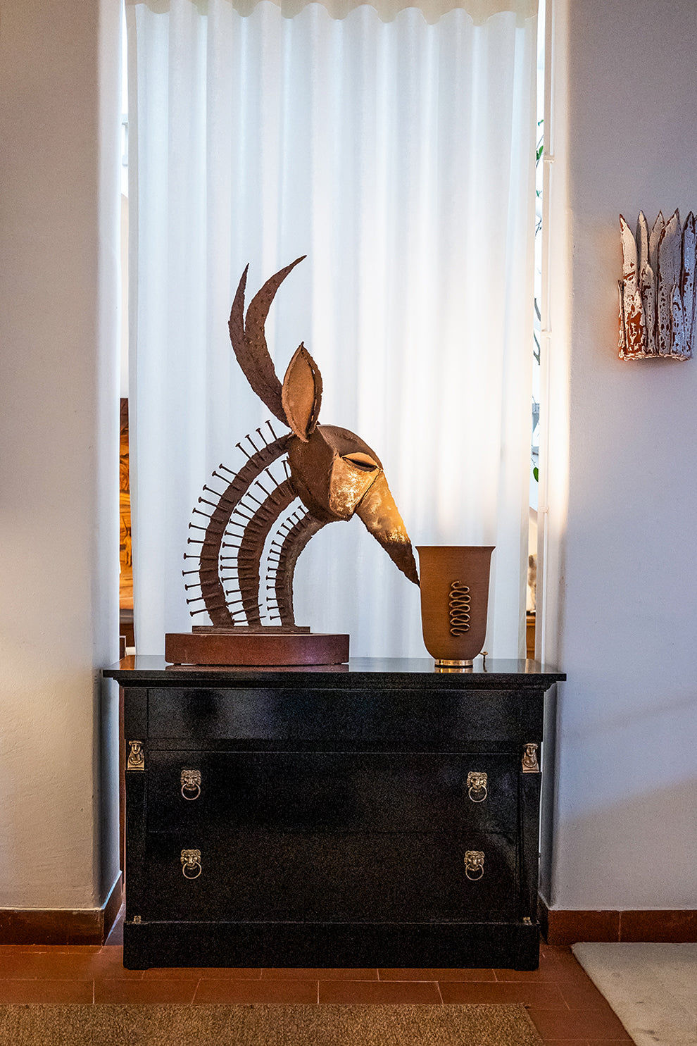 A metal sculpture with tall ears sits on Barracuda Interiors' 19th c 'Return of Egypt' lacquered drawers, next to a brown vase, in front of a white curtain in a well-lit room.