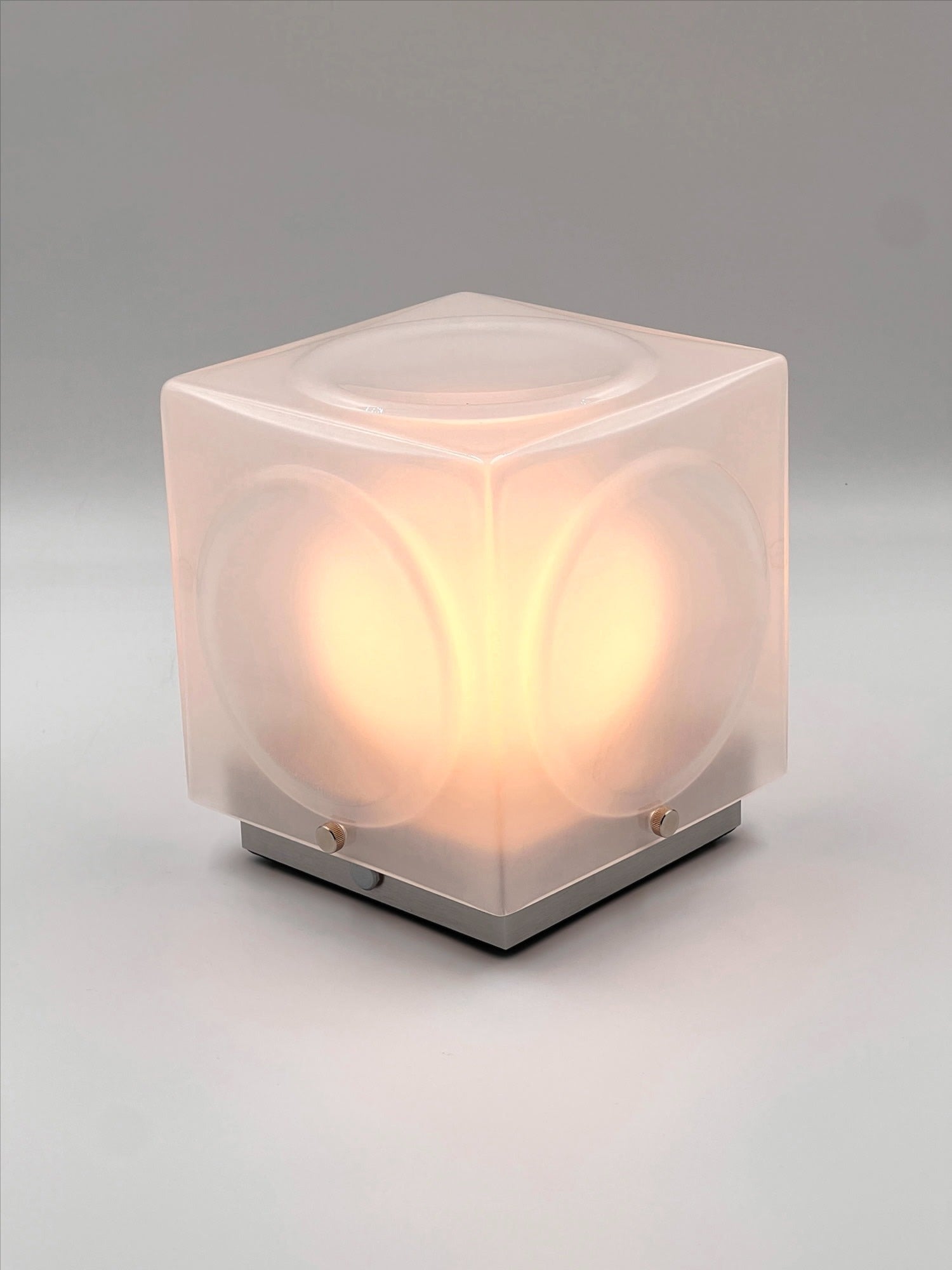 The Chant Portable Table Lamp by Lee Broom features a glowing cube shape, frosted translucent sides, and rounded edges, casting a soft warm light against a plain gray background.