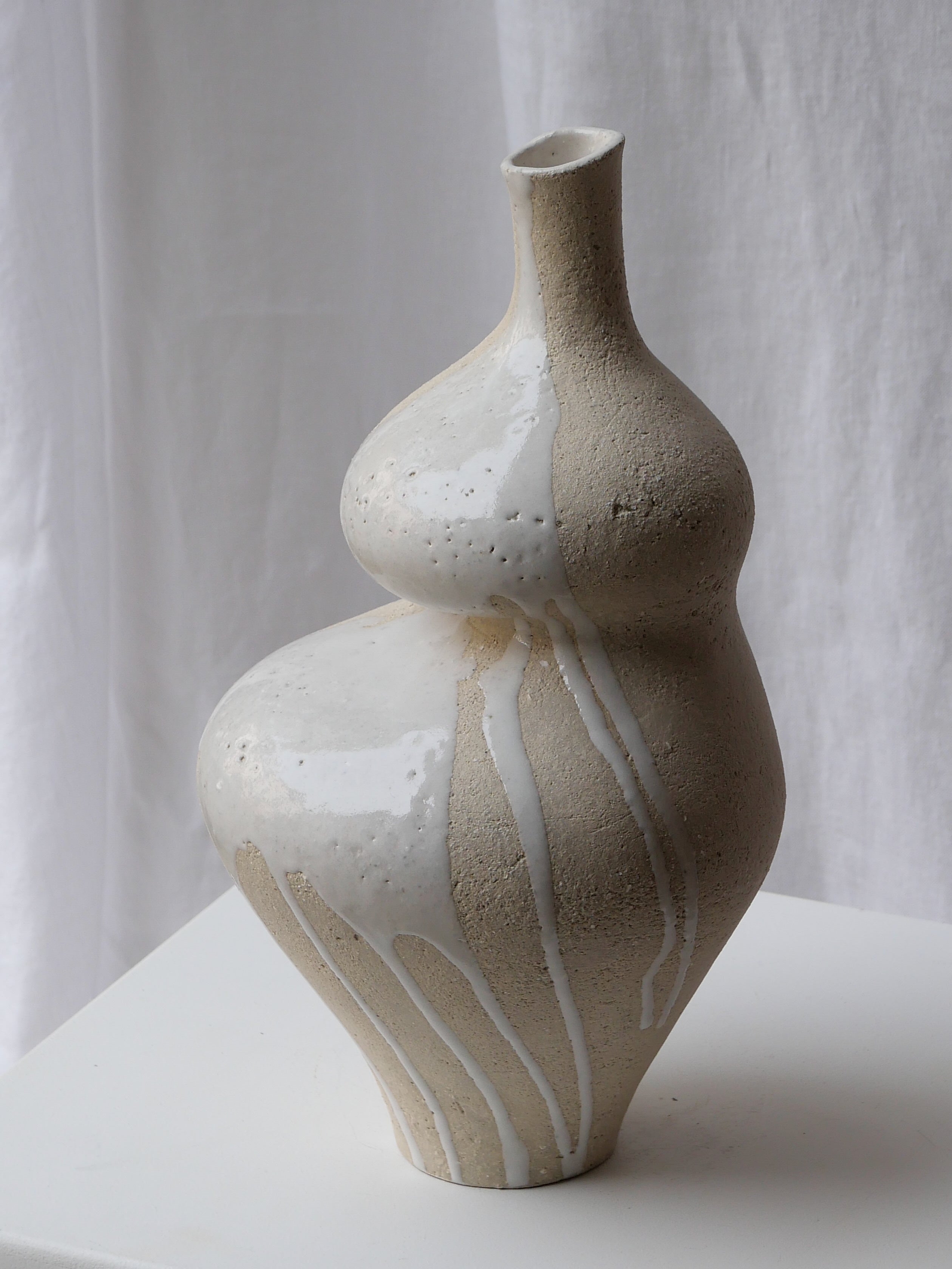 The Lilith White Drip by Lucia Mondadori, a beige handcrafted stoneware vase with a curvy form and glossy white glaze drips, sits on a white pedestal against a soft white fabric backdrop.