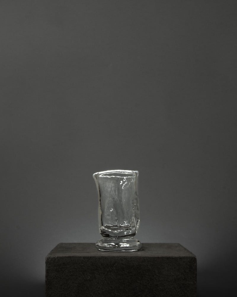 The Organic Shaped Table Glass by Szkło Studio rests on a dark, textured pedestal set against a plain gray background with soft lighting.
