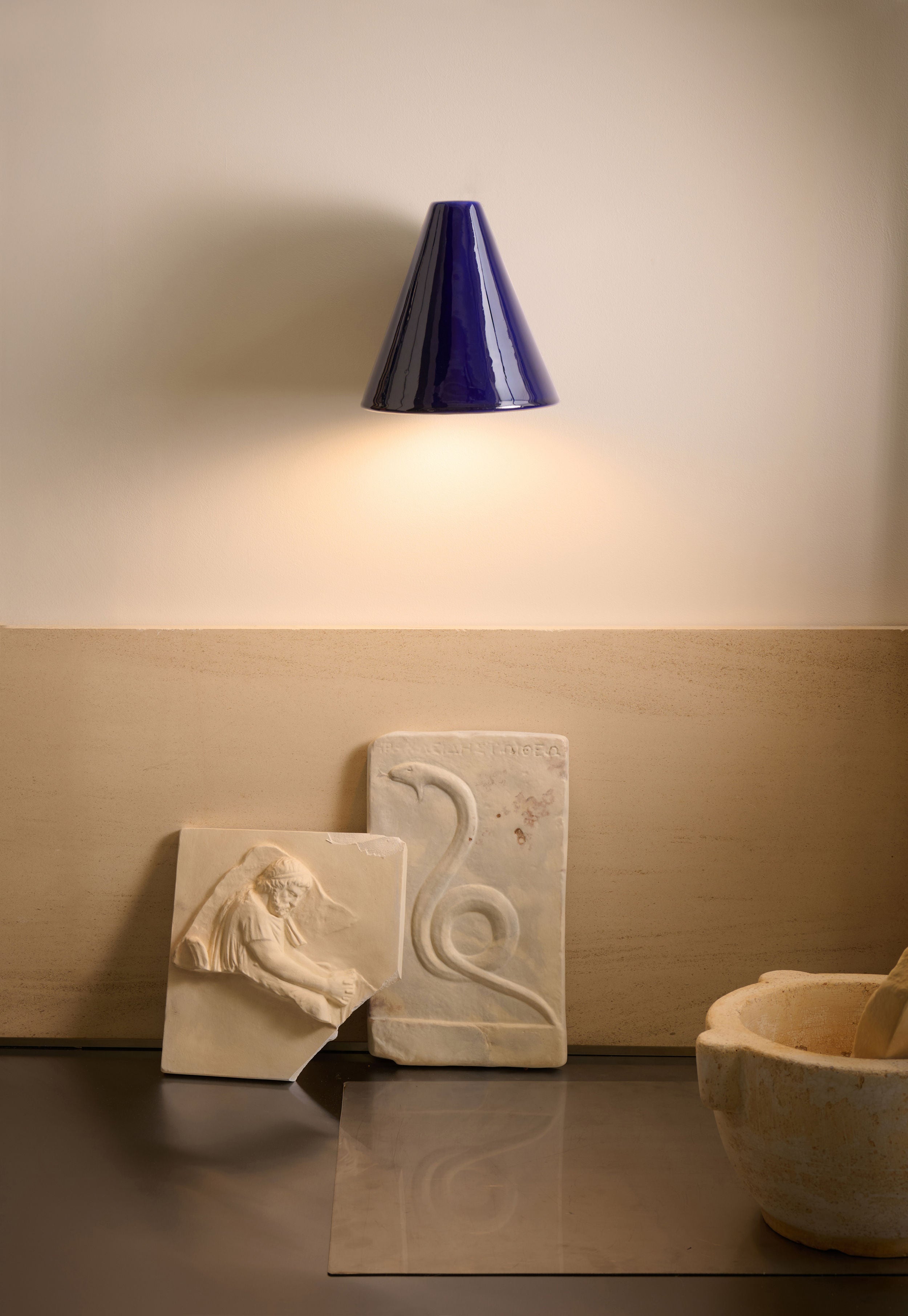 The 'Esoteric' Blue Wall Light by Mariza Galani, cone-shaped in midnight blue, illuminates two stone carvings—a seated figure and a snake motif—on a beige wall, with a handcrafted ceramic bowl partially visible at the lower right.