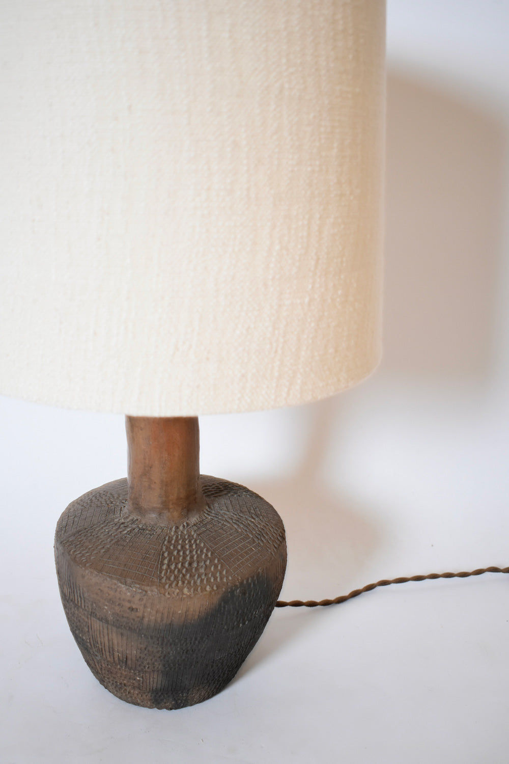 The African Brown Ceramic Table Lamp by Barracuda Interiors (1970s) features a textured dark base and large off-white fabric shade, with the cord visible on a white surface against a white background.