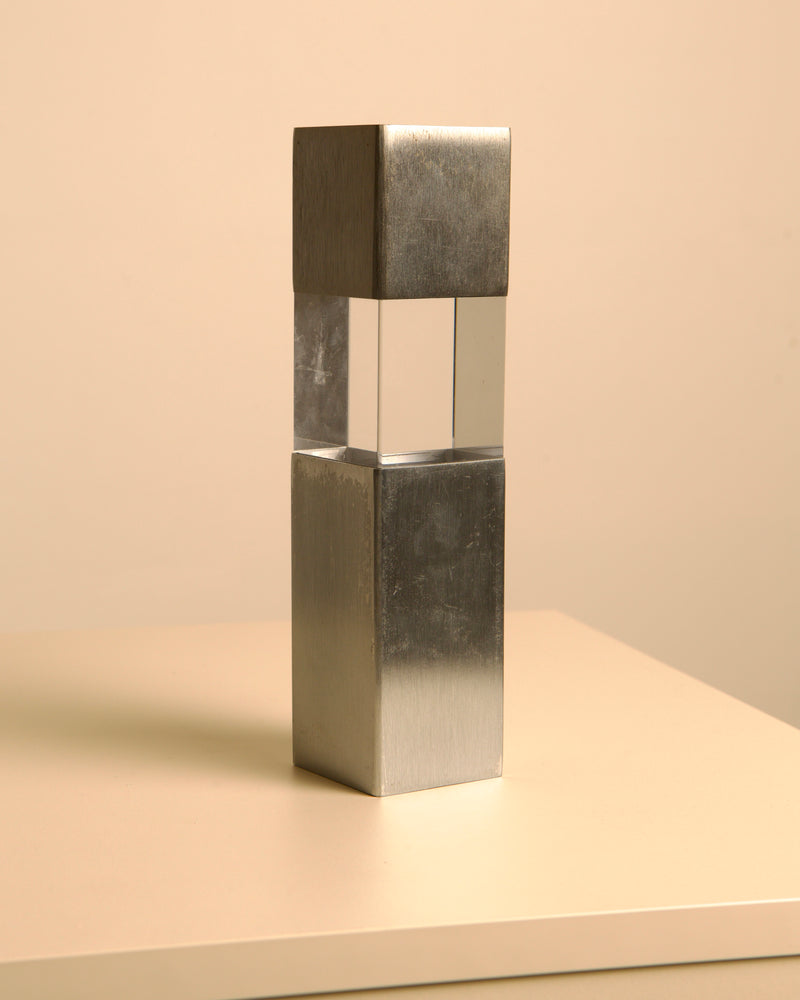 The Treaptyque Italian Sculpture in Metal and Plexiglass 70's features a clear plexiglass cube between brushed metal blocks, standing upright on a beige surface, and evokes classic Italian 1970s design.