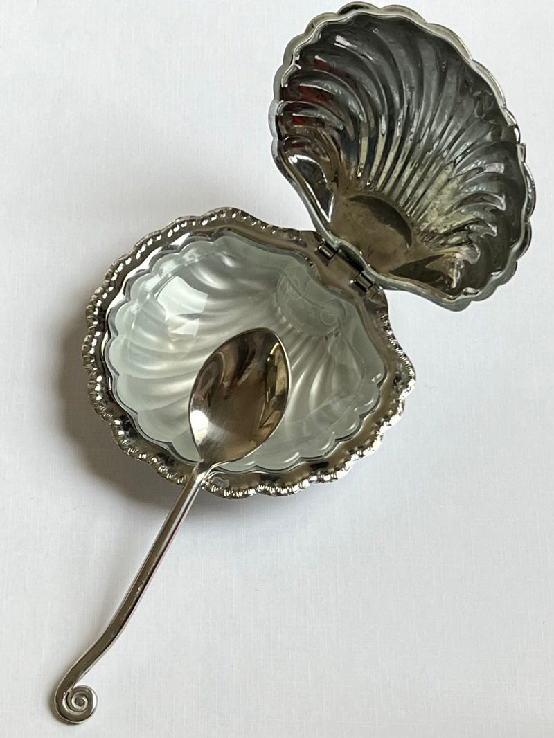 The Manushi Shell Shaped Butter Dish features a hinged lid and matching shell-shaped interior, with an ornate silver spoon inside, all displayed on a white surface.
