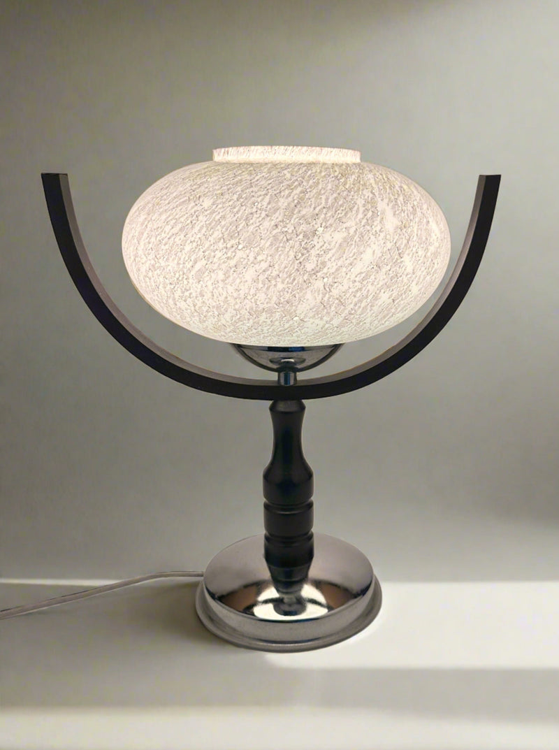The 1960s Sputnik Age Table Lamp by Dodo Vintage features a round, textured white shade on a shiny silver base with two curved black arms. The illuminated lamp rests on a white surface.
