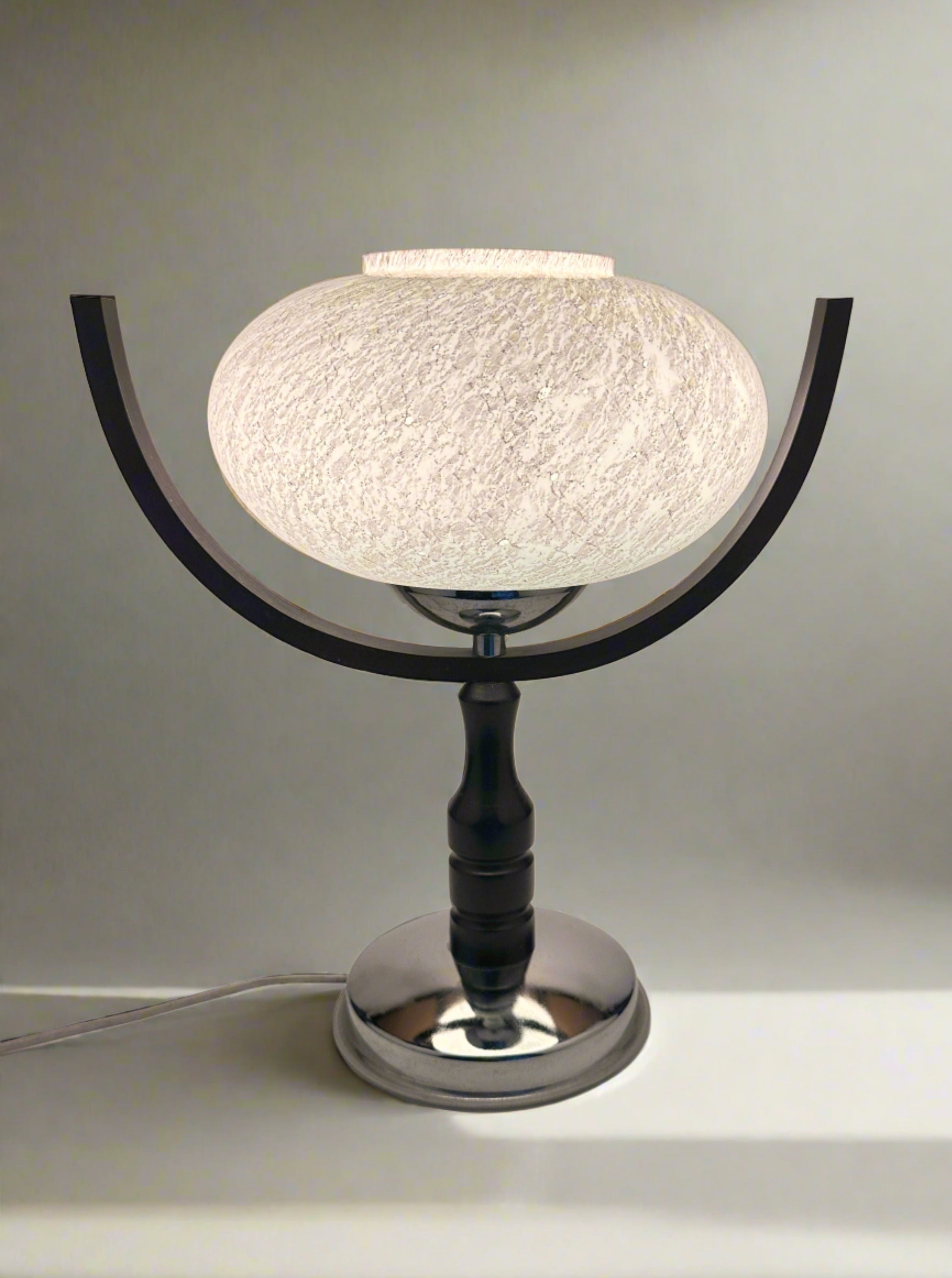 The 1960s Sputnik Age Table Lamp by Dodo Vintage features a round, textured white shade on a shiny silver base with two curved black arms. The illuminated lamp rests on a white surface.