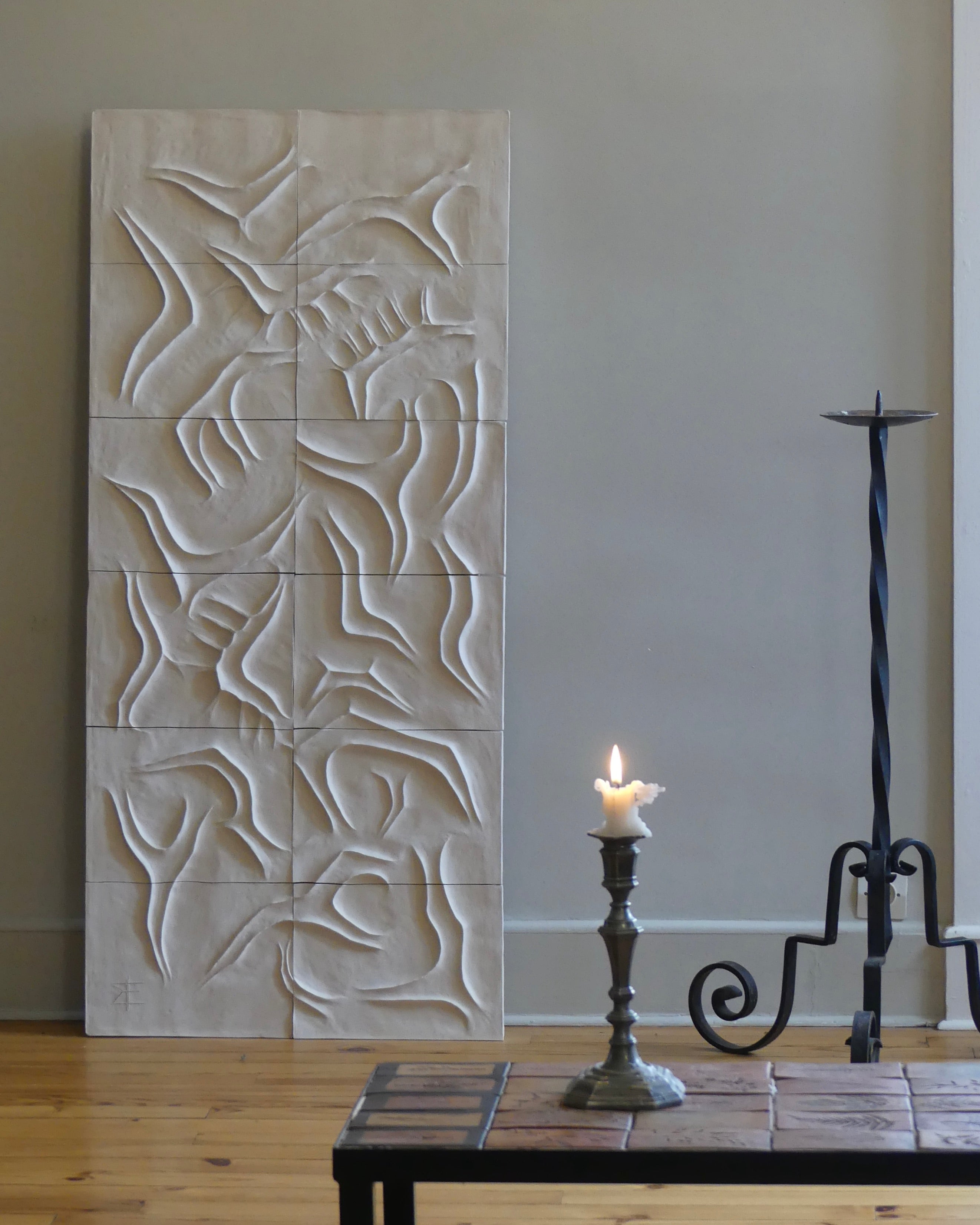 The Scala Dei Turchi wall sculpture by Franck Scala, with abstract flowing patterns, stands against a light gray wall while a small lit candle on a decorative holder near a black iron stand adds to its contemporary charm.