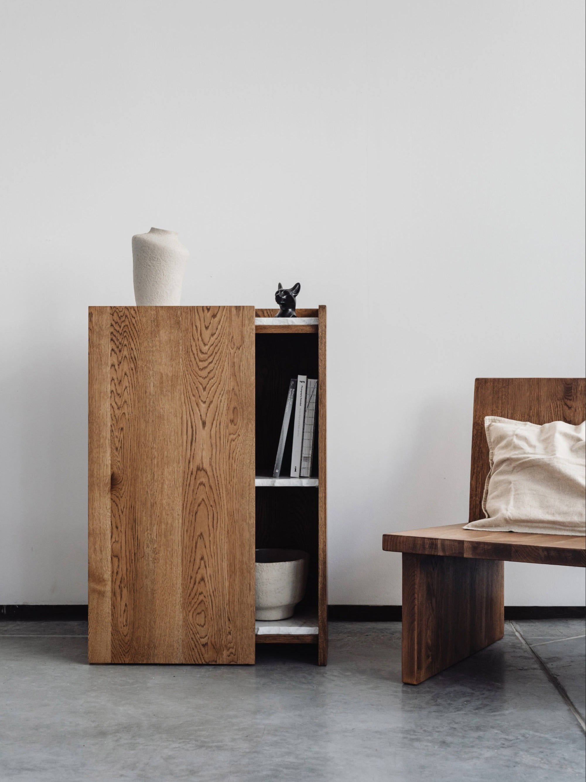 A minimalist interior showcases the un'common PAVO Cabinet, crafted from solid wood with partially open shelves displaying books and a bowl, topped with a vase and black sculpture, beside a wooden bench with a beige pillow against a white wall.