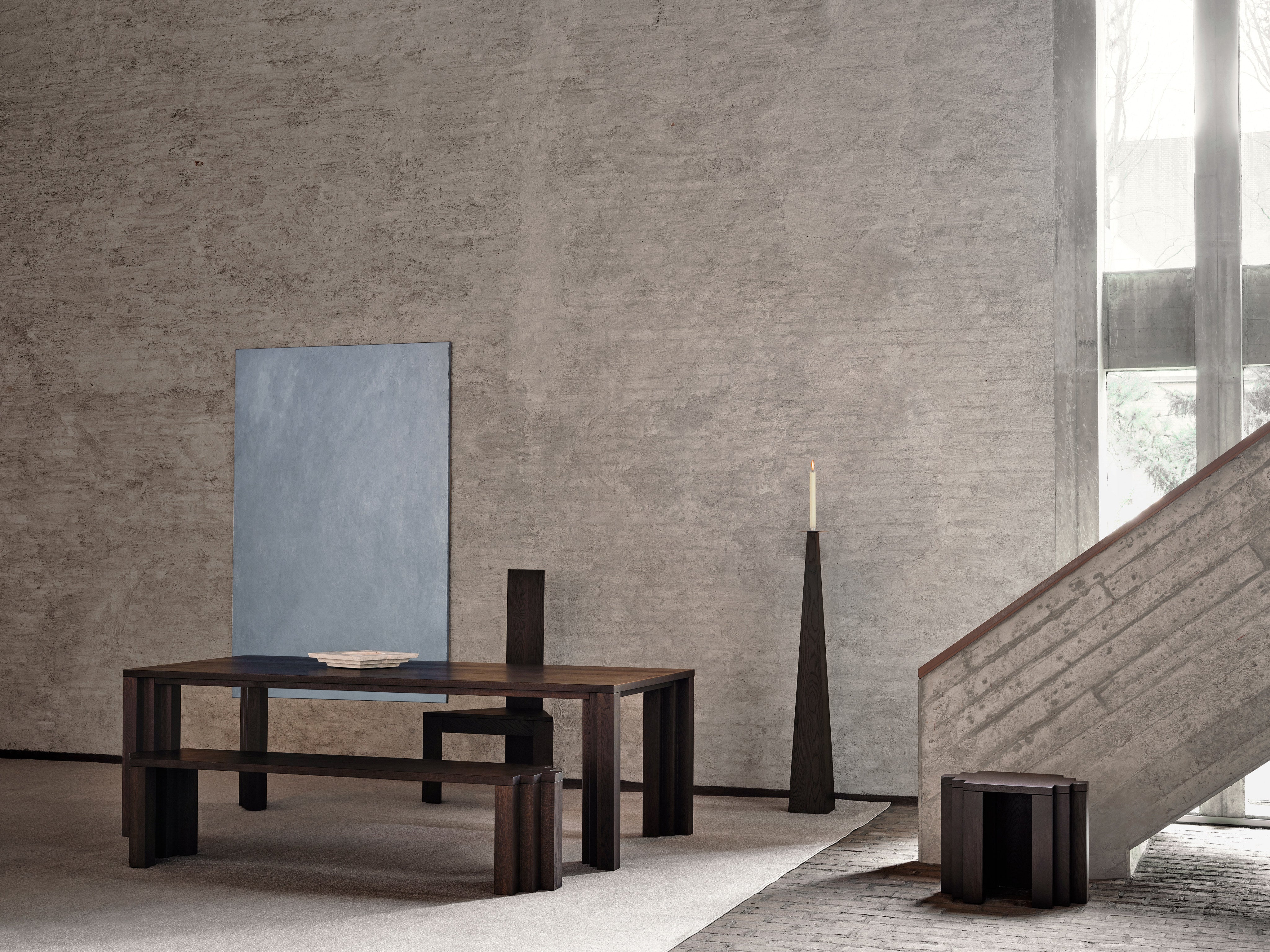A minimalist interior features a dark wooden table, the Mokko Cadence Bench, and a stool on a light rug. Abstract blue art rests by a textured wall, as large windows illuminate customizable solid European oak furniture.