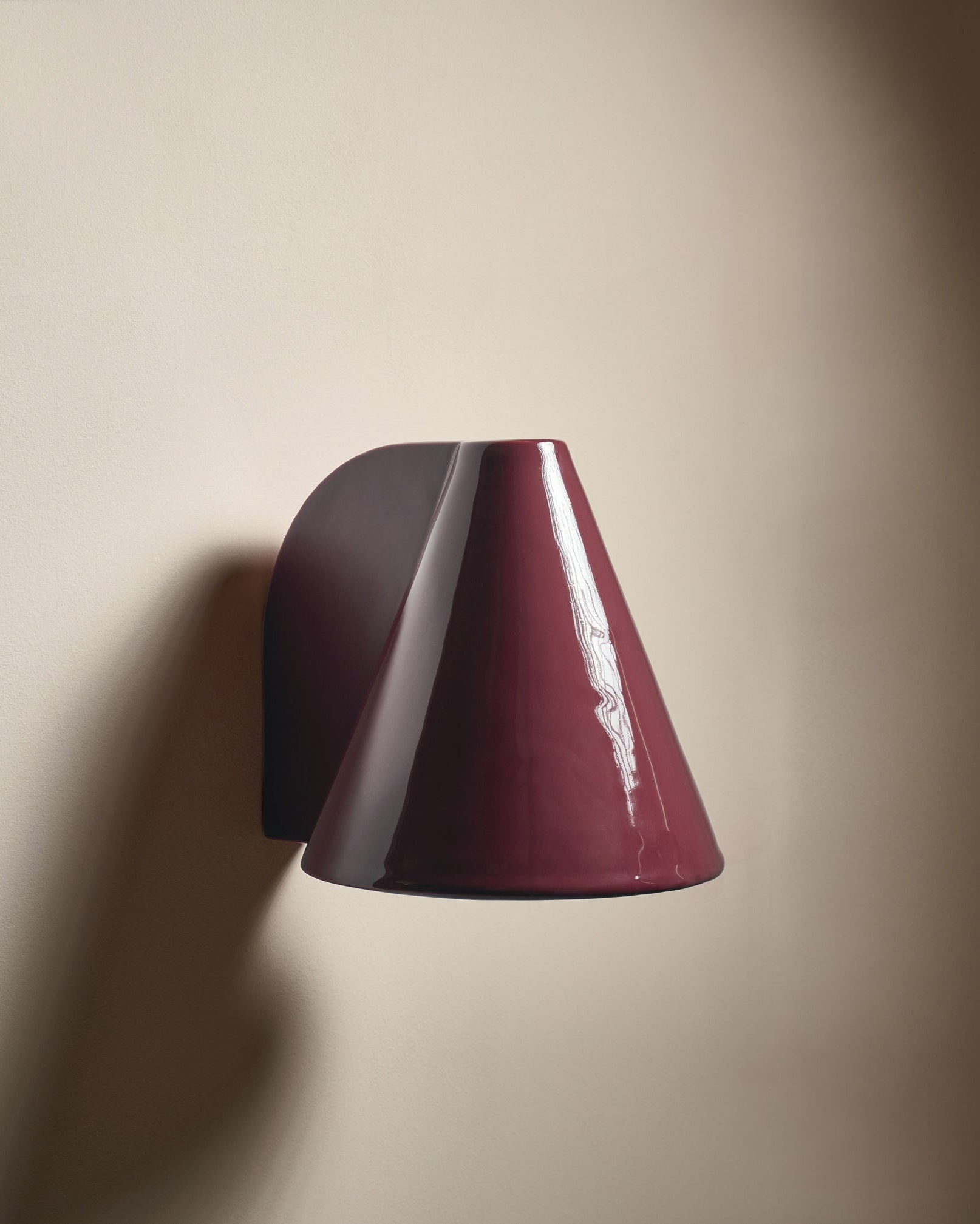 The 'Esoteric' Wall Light by Mariza Galani features a modern, glossy cherry red conical shade that casts soft side shadows when mounted on a smooth off-white wall.