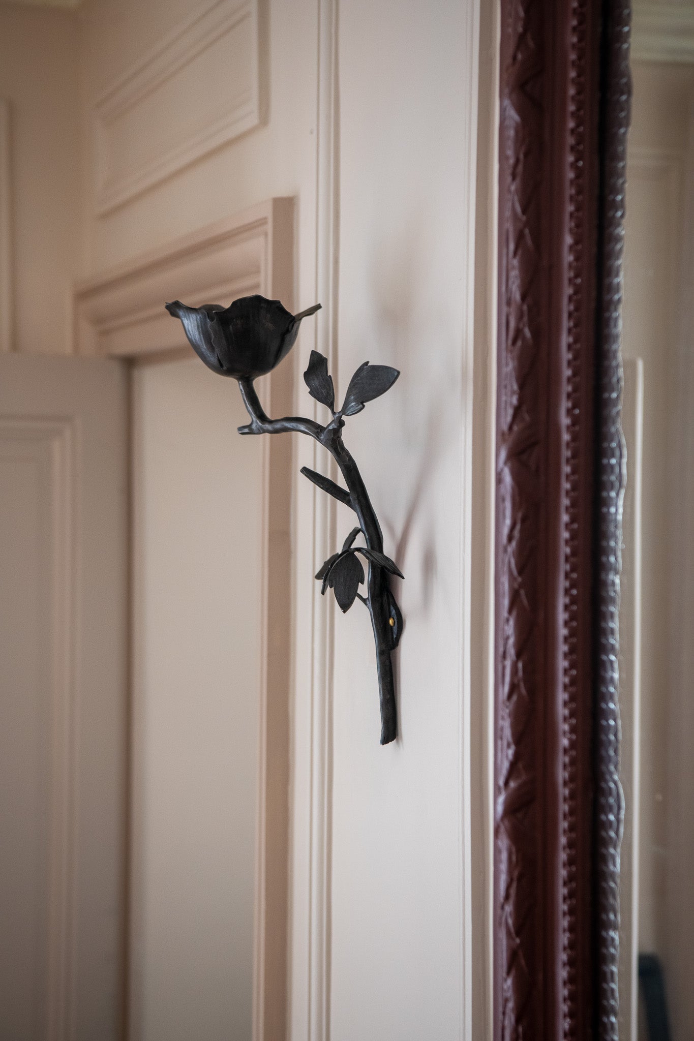 The Clematis Wall Light by Jess Wheeler Studio, a handcrafted forged iron fixture inspired by flowers and branches, is mounted on a light-colored wall next to a large framed mirror in a room with classic molding.
