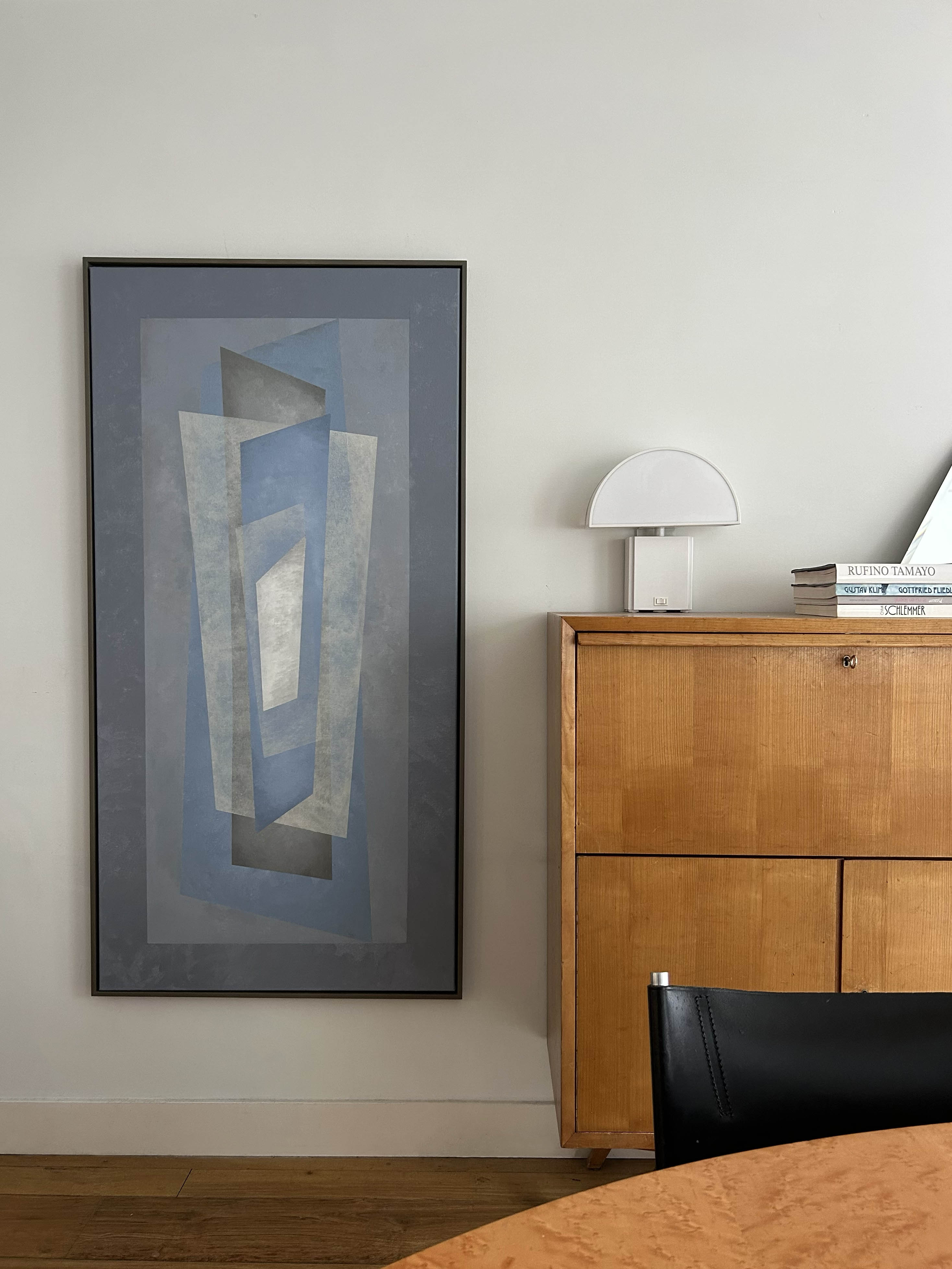 A modern interior showcases the "Floating" Painting by Iris Bavelaar in blue-gray tones, abstract geometric art, a wooden cabinet with books and a white lamp, plus part of a wooden table and black chair in the foreground.
