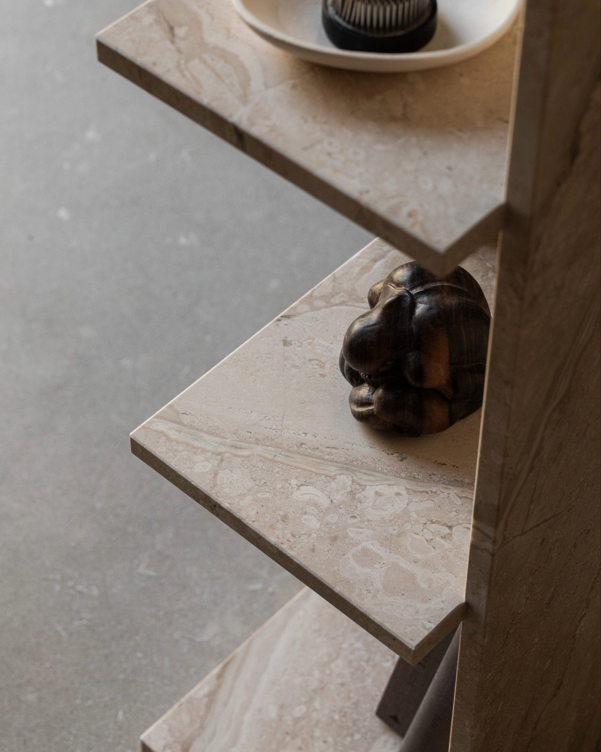 A close-up of the slim CONE Marble Shelf by un'common, featuring three tiers; a small abstract animal sculpture sits on the middle shelf. The gray textured floor in the background highlights this space-saving piece.